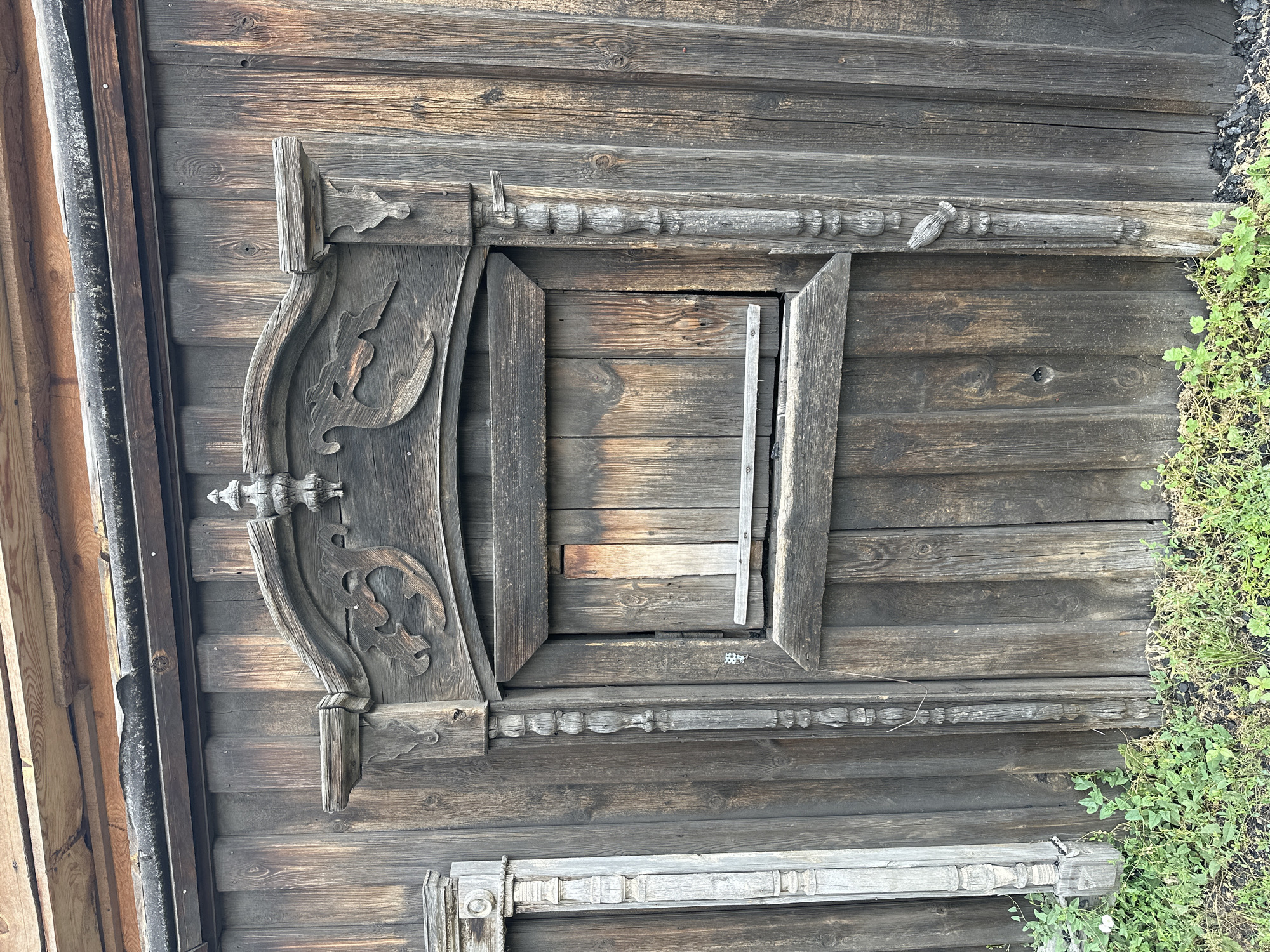 This is a medium shot, eye-level photograph of a weathered wooden structure in the city of Nogornyi, Russia. The central subject is a closed wooden door or window with a decorative wooden frame. The frame is elaborately carved with what appear to be two stylized birds facing each other above a rectangular opening. The wood is dark and aged, showing signs of significant wear and tear, with visible grain patterns and some cracks. There is a horizontal wooden plank positioned across the opening of the central frame. Flanking the central frame are two vertical decorative wooden elements, one to the left and one to the right, both with carved, bead-like segments. To the far left, a portion of another decorative wooden element, possibly a pillar or post, is visible, appearing lighter in color and also showing signs of age. The backdrop is a wall constructed of vertical wooden planks, also weathered and dark. At the bottom of the image, green vegetation, likely grass and small plants, is growing, with some dark debris, possibly coal or ash, mixed in with the soil. The lighting suggests it is daytime, but the sky is not visible.