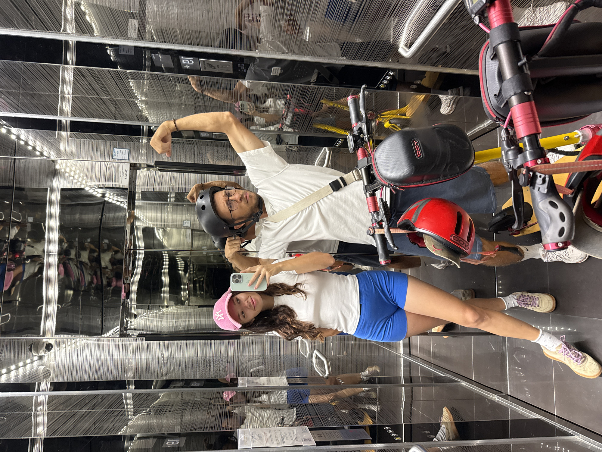 An adult couple stands in a mirrored elevator. The woman, on the left, wears a pink New York Yankees cap, a white t-shirt, and blue shorts. She holds a smartphone in her right hand and is taking a selfie. The man, on the right, wears a black helmet, glasses, and a white t-shirt. He flexes his bicep with his right arm raised. Two electric scooters are positioned in the elevator with the couple. The elevator walls are made of mirrored panels with vertical black lines.