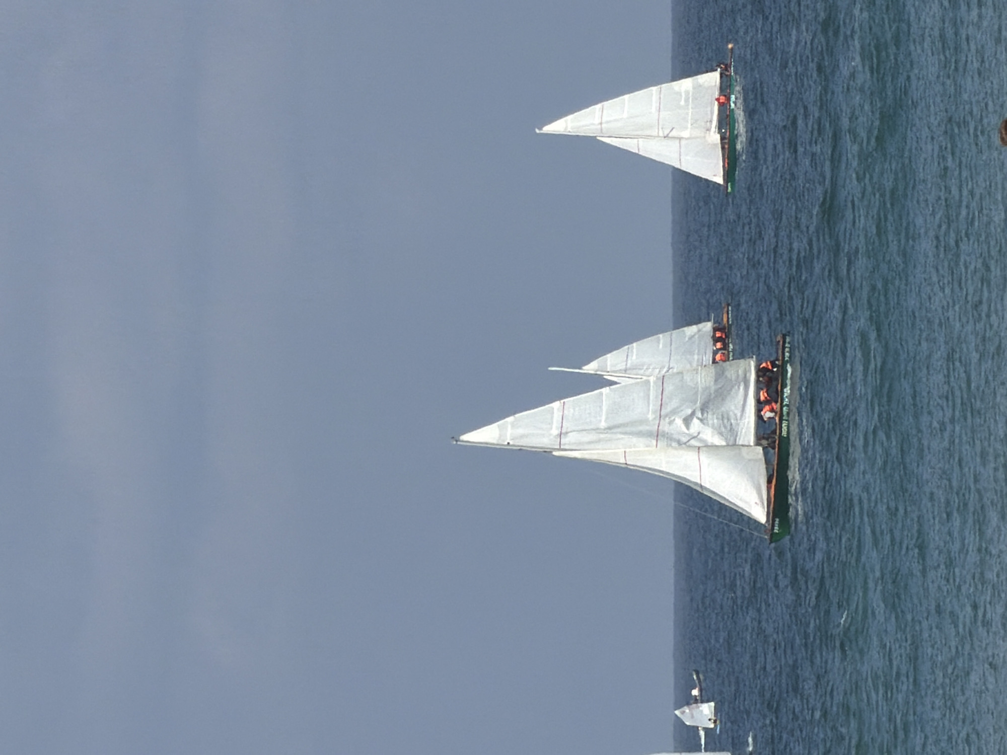 The image shows two sailboats on the Mediterranean Sea off the coast of Tel Aviv, Israel. The sky is overcast. The sailboats are white with green trim, and people wearing orange life vests are visible on the larger boat. The sea is slightly choppy.
