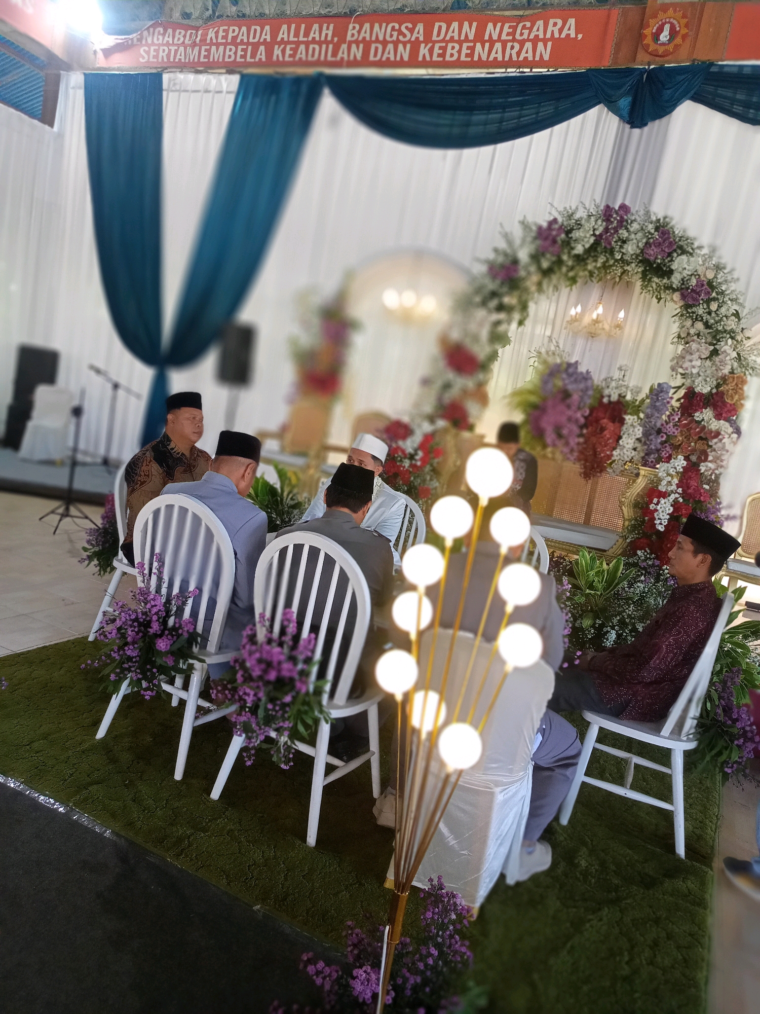 The image depicts an indoor event, likely a ceremony or gathering, with a decorative floral arch as a central feature. Several men, dressed in traditional Islamic attire, are seated on white chairs arranged on a green carpeted surface. The men are wearing peci (caps) and traditional shirts or robes. Some are looking forward, while others are looking down or to the side. Behind the seating area, white drapery is visible, along with teal fabric draped from above, creating a formal backdrop. A banner with text in Indonesian, "MENGABDI KEPADA ALLAH, BANGSA DAN NEGARA, SERTAMEMBELA KEADILAN DAN KEBENARAN," is displayed at the top of the scene. The overall ambiance suggests a solemn and respectful occasion. The mention of Wanadadi, Indonesia, implies the location of this event.