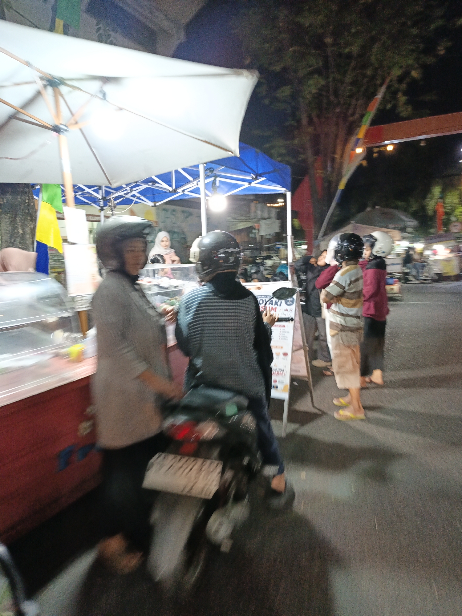 This image depicts an outdoor scene at night, likely a street food vendor or night market in Klaten, Indonesia. In the foreground, a person wearing a grey jacket and a helmet is seated on a motorcycle. To their right, another individual in a striped shirt and helmet is also on a motorcycle. In the background, several other people are visible, some wearing helmets and others without. The scene features food stalls with white and blue awnings, as well as various signs and lights. A tree with foliage is present to the right of the image. The overall lighting suggests it is nighttime, and the presence of motorcycles indicates it is a common mode of transportation in the area.