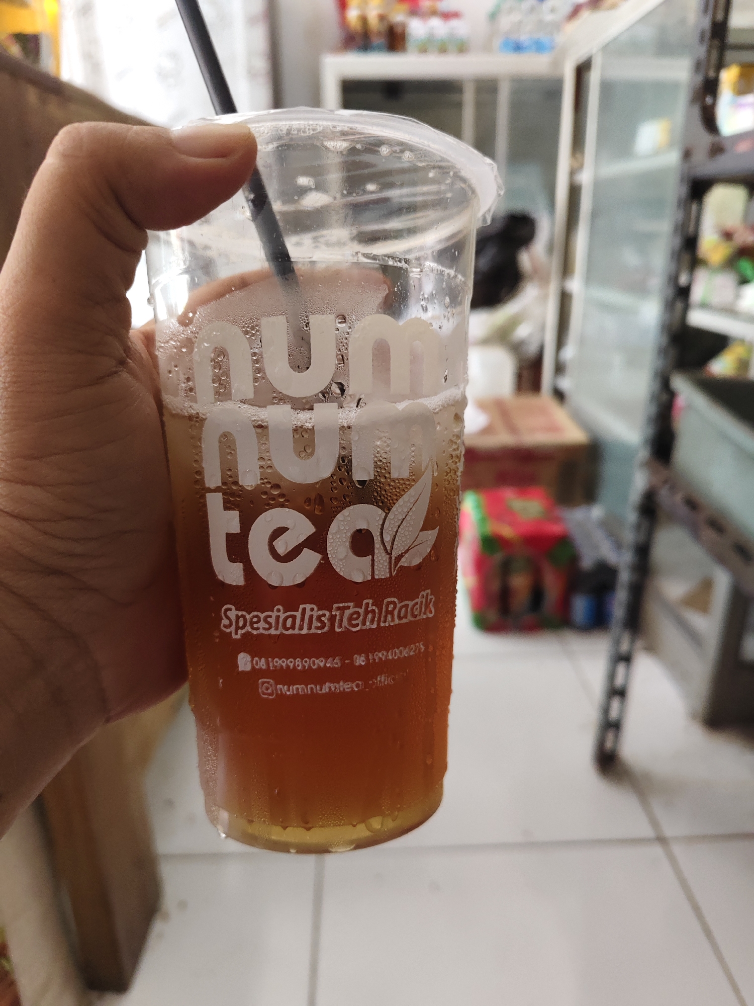 A person's hand is holding a clear plastic cup filled with iced tea. The cup has the logo "num tea" printed in white, and below that, it reads "Spesialis Teh Rack." The drink is amber in color and has condensation on the outside of the cup. A black straw is visible sticking out of the cup. The background is blurred but appears to be an indoor setting with shelves of products. The location is identified as Bangsalsari, Indonesia.