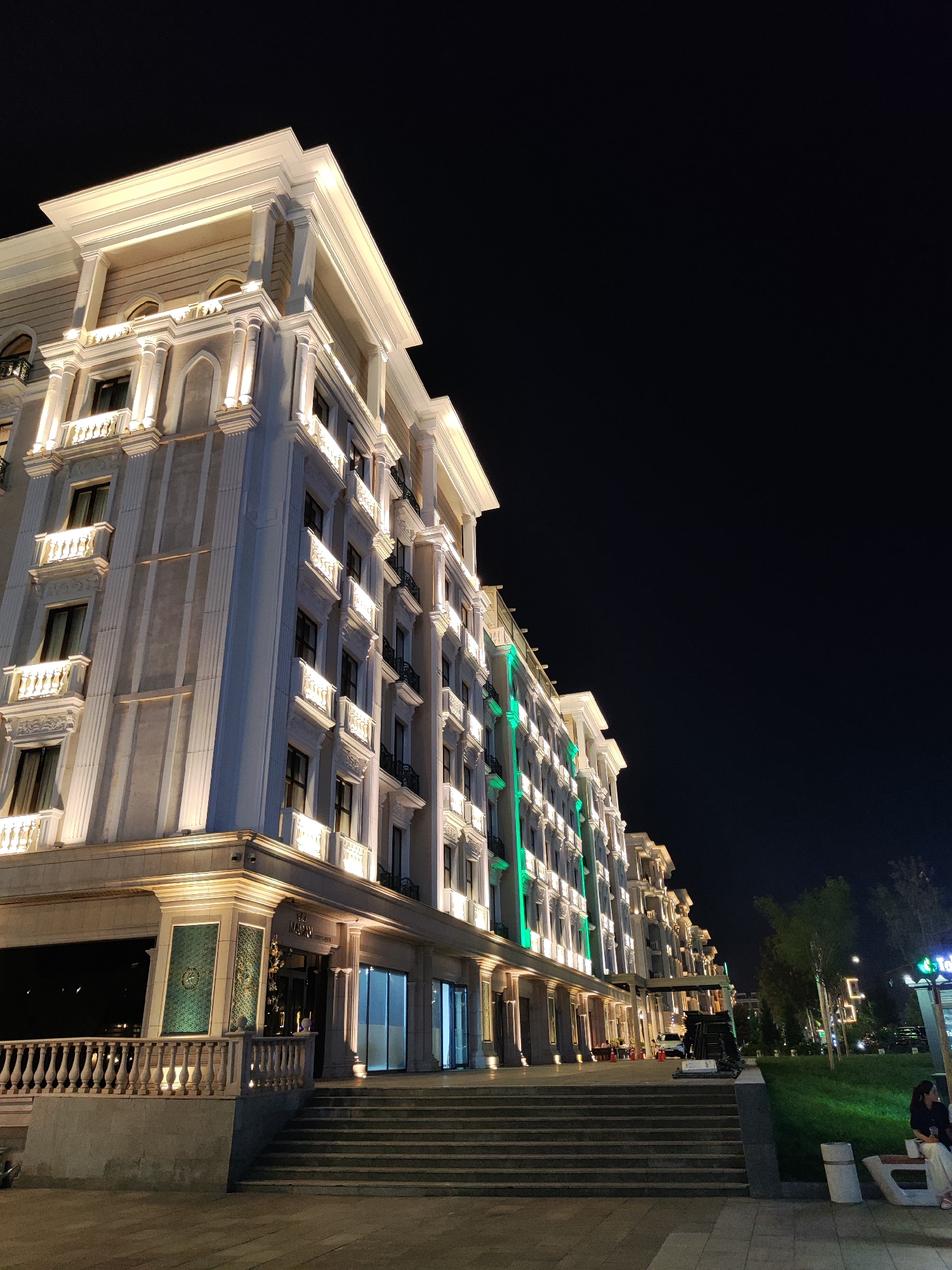 A night time, low angle exterior shot of a grand, multi-story building with classical architectural elements, possibly a hotel or a luxury residential building. The building is illuminated with warm, golden lights on the facade and green lights accentuating the upper floors. The facade features ornate columns, balconies with decorative railings, and arched windows. In the foreground, a wide set of stone stairs leads up to the building entrance, which is flanked by glass doors and decorative green panels. A white railing runs along the side of the stairs. To the right of the building, a well-lit green lawn area is visible, with trees and streetlights. A few people are seated on a bench in the background. The overall impression is one of elegance and sophistication. Based on the architectural style and the presence of greenery and streetlights, this scene is consistent with an area like the Tashkent City International Business Center in Tashkent, Uzbekistan, specifically near buildings that were part of the initial development phases, like the aforementioned Hilton Tashkent City.