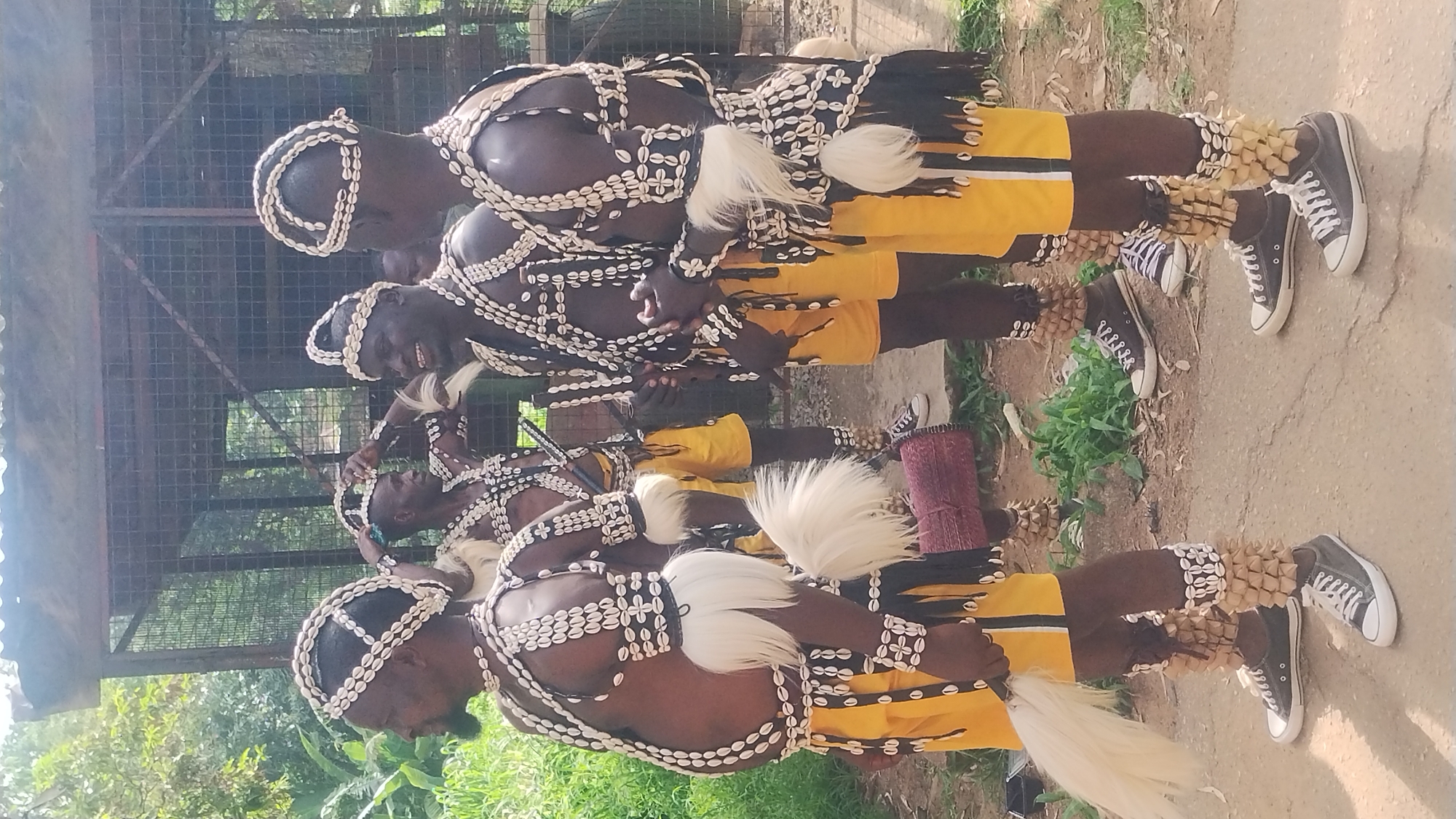 This photograph depicts four men dressed in traditional attire, adorned with cowrie shells and white feather decorations. They are standing on a dirt path in what appears to be an outdoor, possibly rural, setting. Behind them, a metal cage structure is visible, overgrown with vegetation. One man on the far left holds a white feather whisk. The men are wearing yellow shorts and dark sneakers. The man second from the left is smiling broadly and adjusting his headdress. The image captures a moment of cultural expression, possibly during a festival or ceremony. The natural lighting suggests it was taken during the daytime. The presence of the cage structure could indicate a zoo, an enclosure for animals, or possibly a part of a cultural performance space.