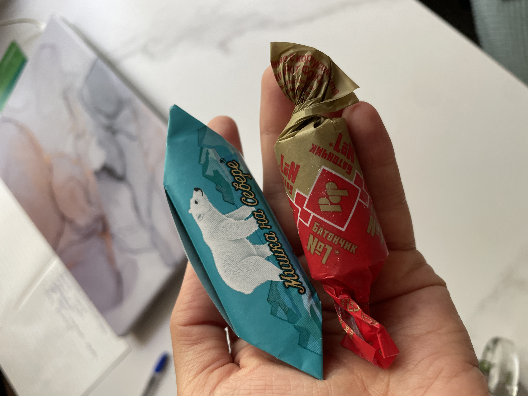 A hand is holding two candies. The top candy is wrapped in teal foil with a white polar bear and mountains depicted on it, along with text in Russian that reads "Мишка на Севере" (Bear in the North). The bottom candy is wrapped in red and gold foil. The red foil has some text and a diamond shape, and the gold foil has Russian text that appears to read "Батончик №1" (Bar No. 1). The background shows a notebook with a marble-like pattern and a blue pen. The scene is likely indoors, on a white surface, possibly a table. The location in Zheleznogorsk, Russia is not discernible from the image.