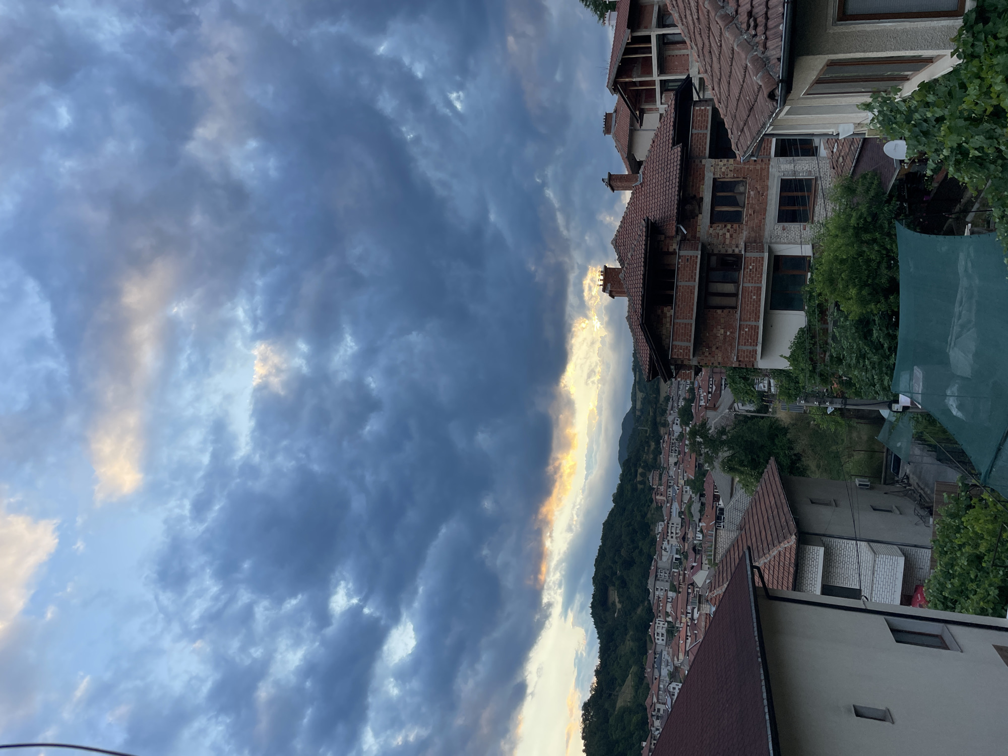 The image displays a view of Zlatograd, Bulgaria, characterized by its distinctive architecture and mountainous terrain. The foreground is dominated by the rooftops of several houses, their red-tiled roofs a prominent feature. These houses exhibit a mix of styles, with some appearing to be older, traditional structures and others in various stages of construction, revealing exposed brickwork and concrete.
Beyond the immediate residential area, the town of Zlatograd unfolds across a gentle slope, showcasing a dense cluster of buildings with similar red-tiled roofs. The urban landscape gradually merges into the lush green foothills of the Rhodope Mountains, which form a dramatic backdrop to the town. The mountains are densely covered with trees, indicating a natural, less developed area surrounding the settlement.
The sky above is a spectacle of dramatic cloud formations. Dark, heavy clouds dominate the upper portion of the image, suggesting an approaching storm or the aftermath of one. However, towards the horizon, a break in the clouds reveals a sliver of bright, warm light, possibly the setting sun, casting a golden and orange glow that contrasts sharply with the cooler tones of the clouds and the surrounding landscape. This interplay of light and shadow adds a sense of depth and atmosphere to the scene.
The image captures a moment of transition, where the rugged beauty of the natural environment meets the human settlement. The presence of construction suggests ongoing development within the town. No people are visible in the frame, focusing the attention on the architectural and natural elements of Zlatograd.