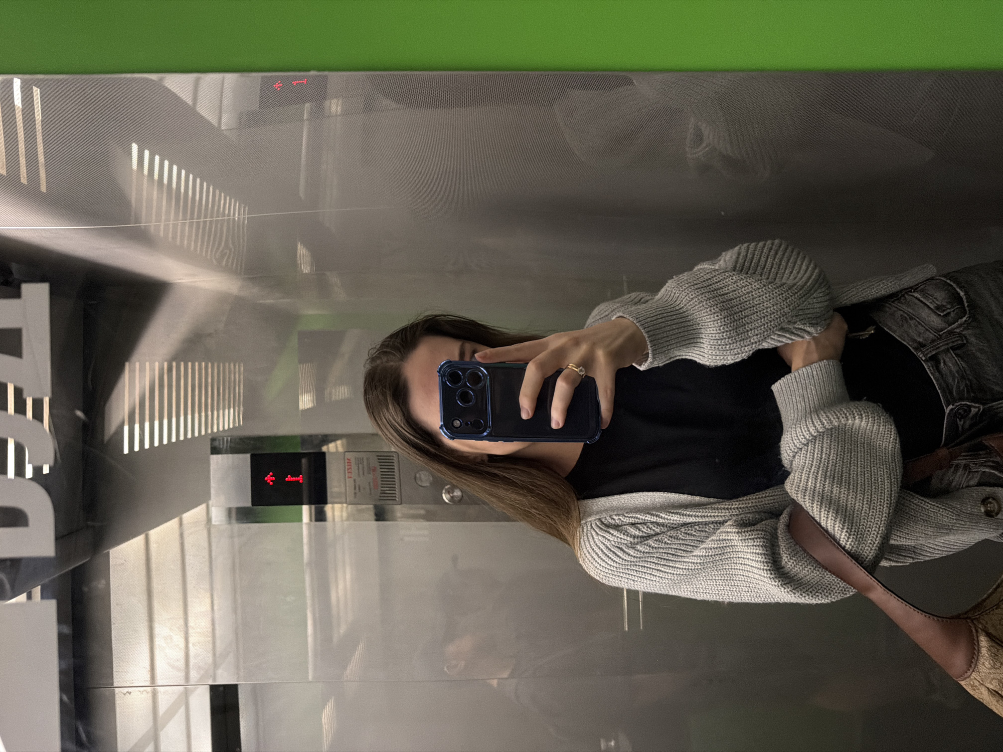 A young woman with long brown hair takes a selfie in an elevator. She is wearing a light grey cardigan over a black top and dark grey jeans. She holds a black smartphone in her right hand, covering her face. A gold ring with a single stone is visible on her right ring finger. A brown purse with a patterned strap is slung over her left shoulder. The elevator walls are reflective stainless steel, with horizontal lines creating a textured pattern. A green panel is visible on the right side of the elevator. A red indicator light showing an upward arrow and the number "1" is visible on the elevator wall. In the reflection of the elevator door, a man wearing a dark baseball cap can be seen. The elevator appears to be located in a building in Kyiv, Ukraine, based on the signage visible in the reflection, which includes Cyrillic lettering.
