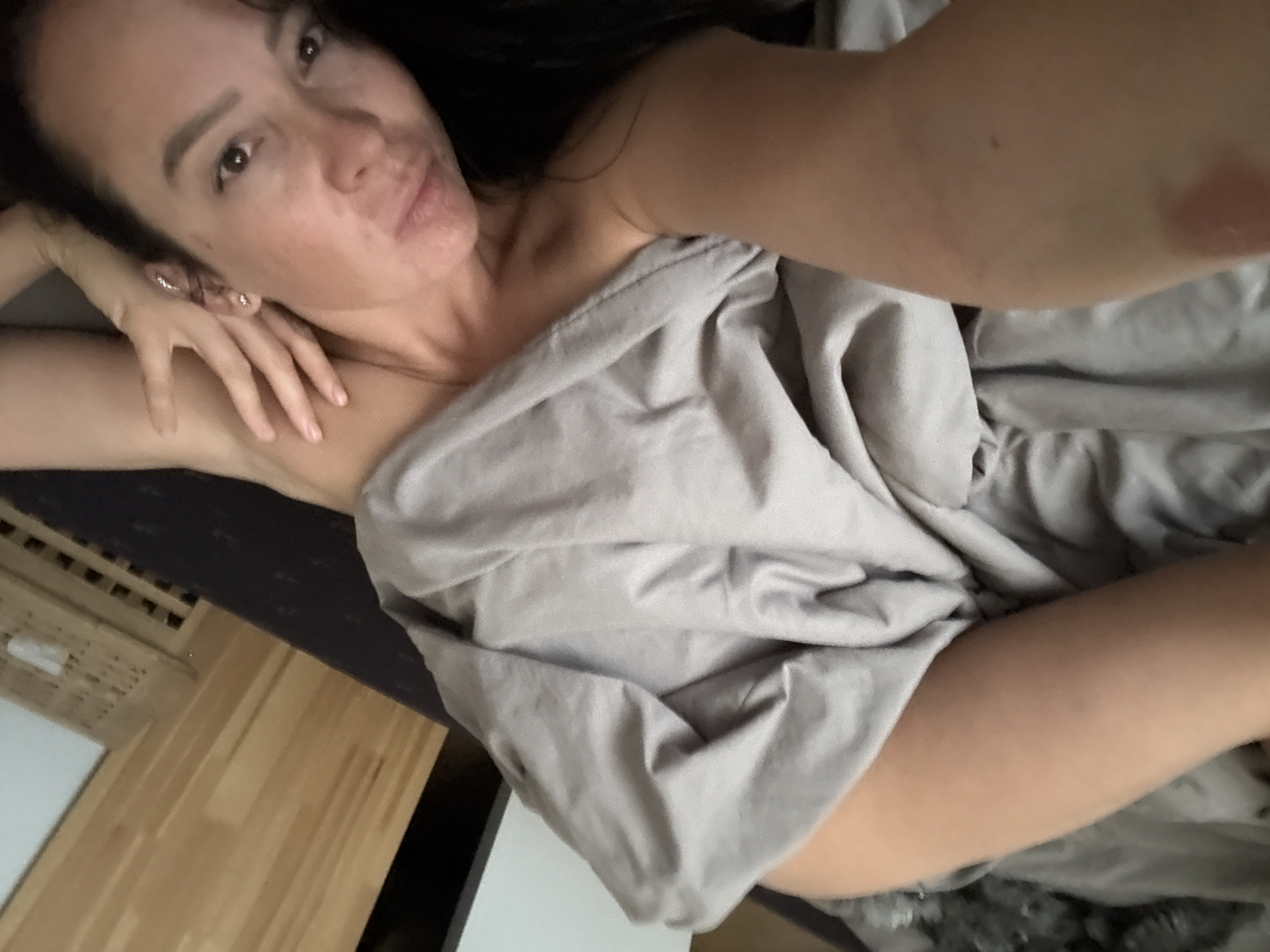 A woman is lying in bed, covered by a gray blanket. She is holding her hand up near her face. The image may have been taken in Chaikovsky, Russia. The environment includes a wooden headboard and flooring.