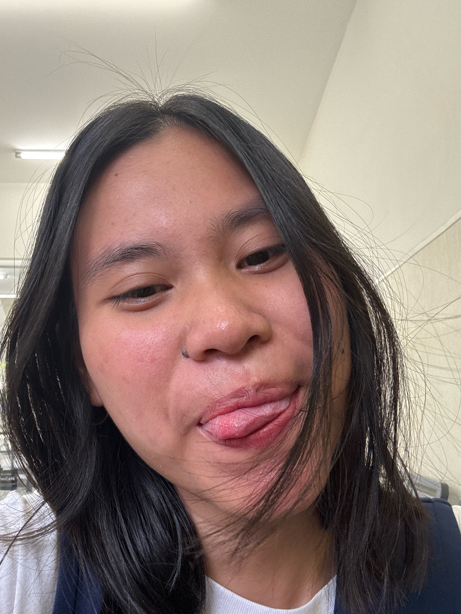 A close-up, low-angle, front-facing selfie of a person with dark hair, sticking out their tongue and making a playful face. The person's hair is long and black, with some stray strands framing their face. They have a small nose piercing. The background is a plain, lightly colored wall with fluorescent lighting visible. The setting appears to be an indoor environment, possibly a classroom or office space. This image was captured on October 30, 2023, at 7:14 AM, within the city of East Jakarta, Indonesia.