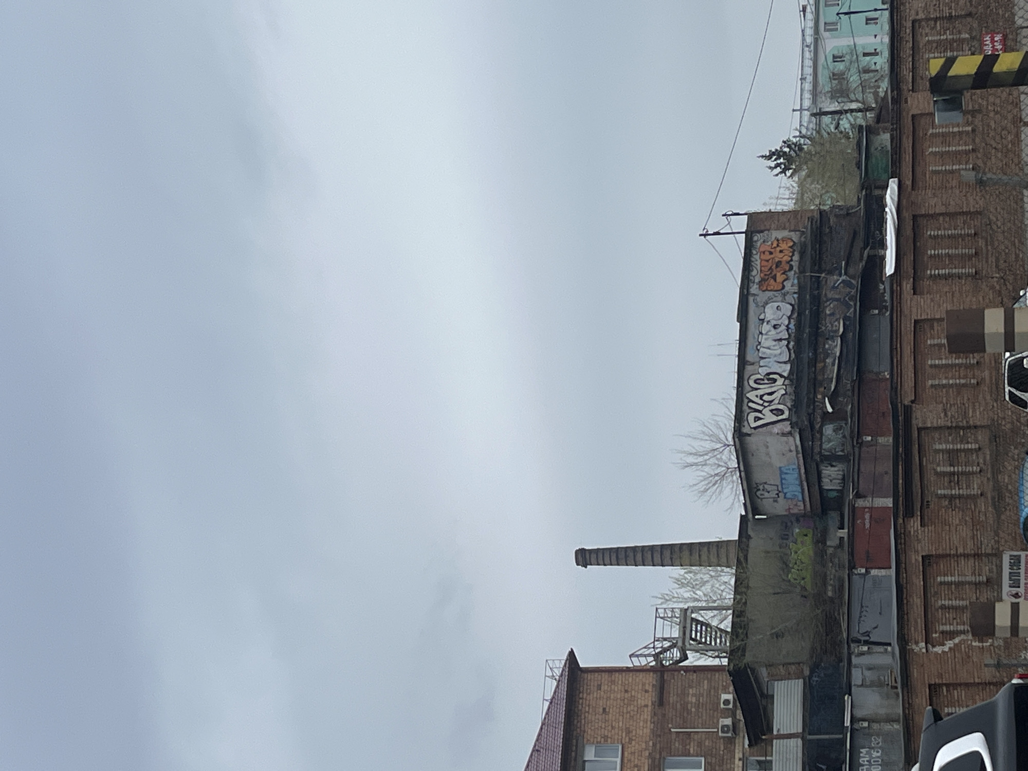 The image depicts a cloudy urban scene in Krasnoyarsk, Russia. A brick building with a chimney is visible, along with adjacent structures showing signs of age and disrepair. Graffiti is present on one of the buildings. Overhead power lines stretch across the sky. The scene suggests an industrial or older part of the city.