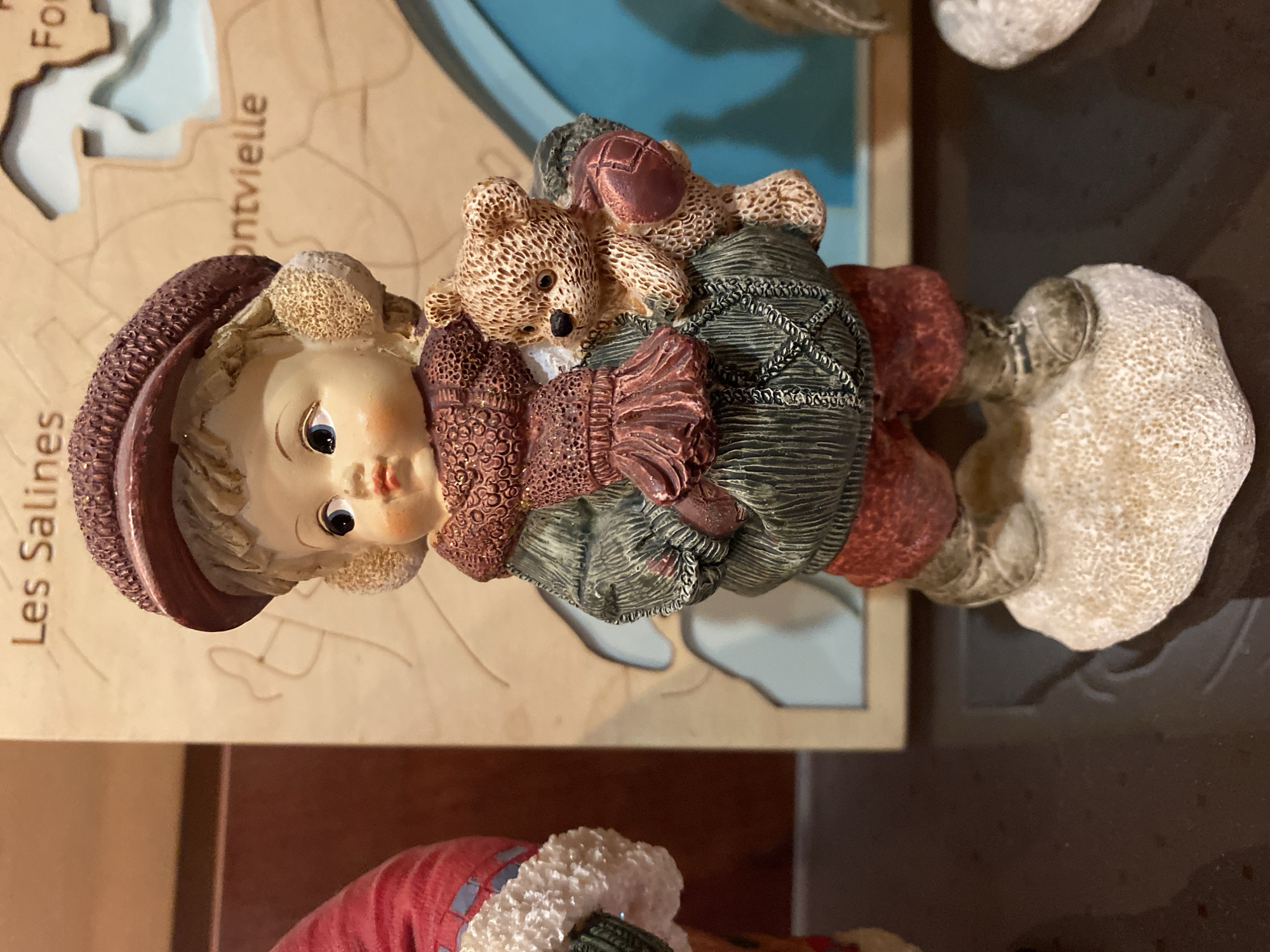 This image features a figurine of a child holding two teddy bears. The figurine is light-skinned with rosy cheeks and blue eyes, wearing a burgundy hat and a green coat with red accents. One teddy bear is brown and the other is also brown with darker brown stitching. The figurine is positioned on a wooden surface with a map-like design. Visible text on the map includes "Les Salines" and "montvielle". The background is a dark brown surface. The lighting appears to be indoor, with no discernible indication of time of day or weather. The setting seems to be a decorative display or shelf.