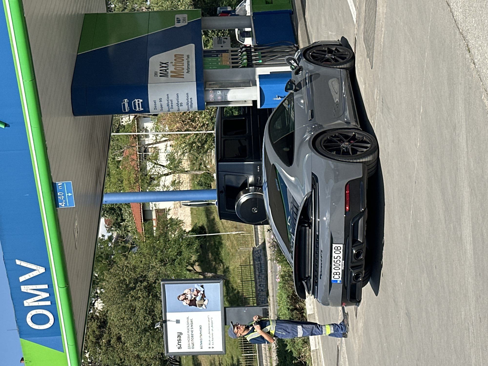 This image depicts a scene at an OMV gas station in Veliko Tarnovo, Bulgaria, during daylight hours. A gray Porsche 911 GTS, bearing the license plate "CB 0055 OB," is parked at a fuel pump. A gas station attendant, wearing a blue uniform with reflective stripes and a cap, stands to the left of the vehicle, facing away from the camera and appearing to be engaged in a task related to his job. In the background, a black Mercedes-Benz G-Wagon is also visible near the fuel pumps. Above the pumps, a digital sign displays "4.40." To the left, a billboard advertises "sinsay" with text in Bulgarian. The environment includes trees, buildings, and a paved road surface.