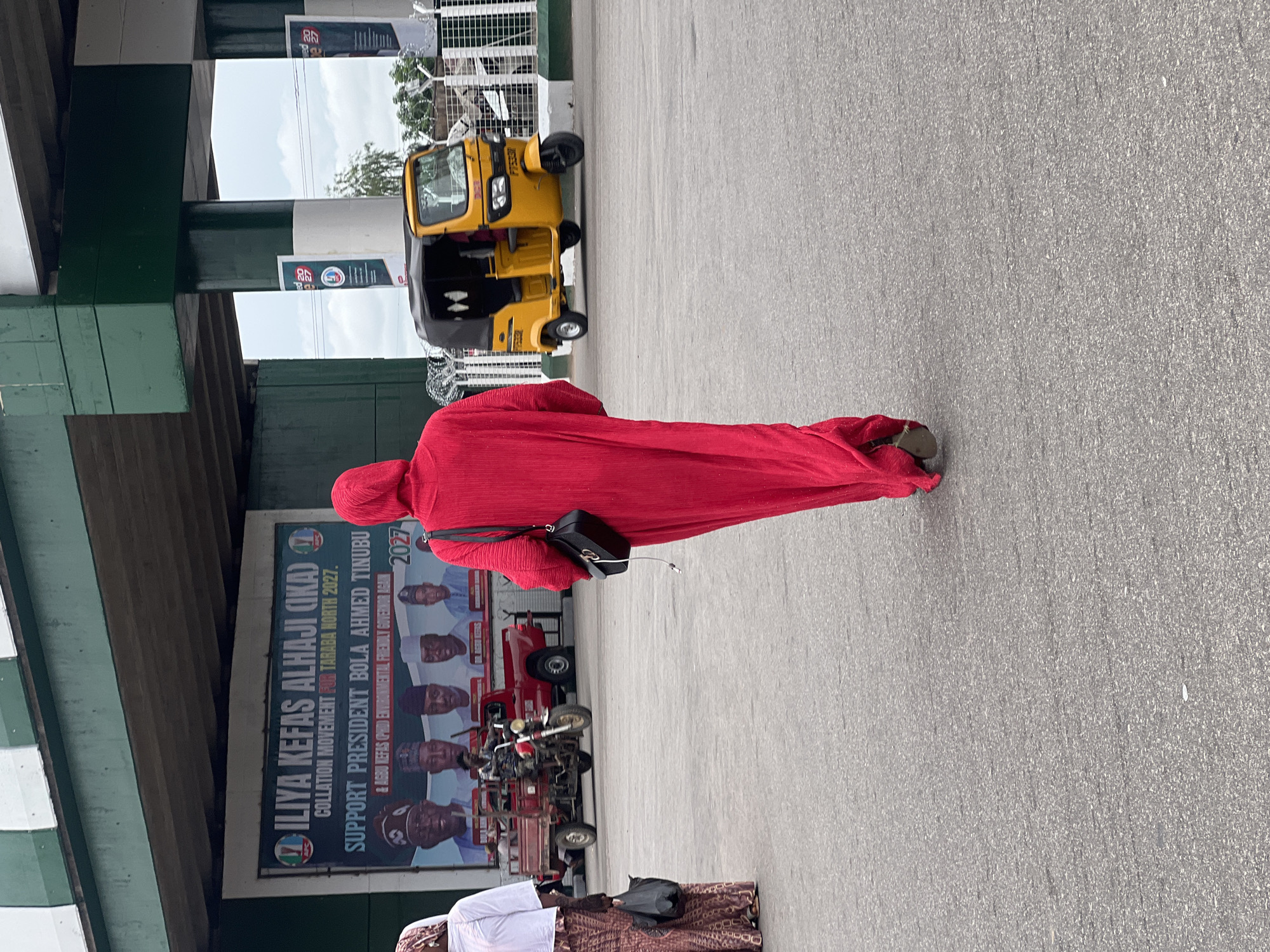 An overhead shot captures a scene in Jalingo, Nigeria, under a green and white overpass. A person in a long, flowing red garment walks away from the viewer toward a large banner on a building. The banner, partially obscured by the red-clad figure, features portraits of men, the words "ILIYA KEFAS ALHAJI (IKA)" and "COLLATION MOVEMENT FOR TARABA NORTH 2027," and "SUPPORT PRESIDENT BOLA AHMED TINUBU & AGWU KEFAS (PHC) ENVIRONMENTAL FRIENDLY GOVERNOR AGAIN 2027-". To the right, a bright yellow, three-wheeled auto-rickshaw, known as a "Keke Napep," is parked. In the background, a red utility truck is also visible, along with barbed wire fencing and trees. Another person in a white top and patterned skirt stands to the left of the frame, carrying a black bag. The ground is paved asphalt.