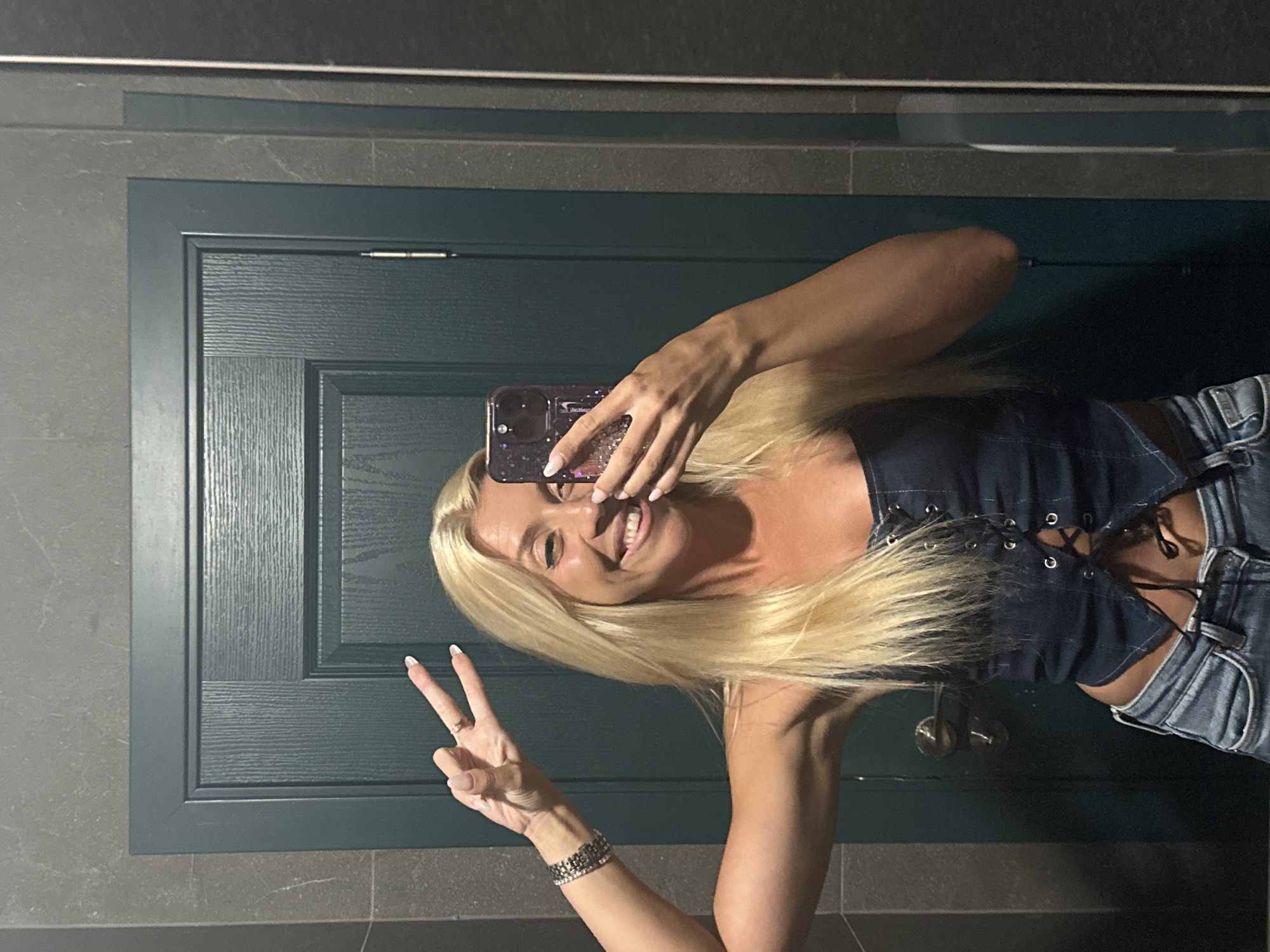 A blonde woman is taking a selfie in a bathroom, wearing a denim corset top and jeans. She is holding her phone in her right hand and making a peace sign with her left hand. The background shows a dark teal door with vertical paneling and a silver doorknob. The walls are tiled with grey stone. The image is well-lit, suggesting artificial lighting. The location is a bathroom, likely in a public establishment or a home. There are no discernible elements in the image that would indicate the city of Pazardzhik, Bulgaria.