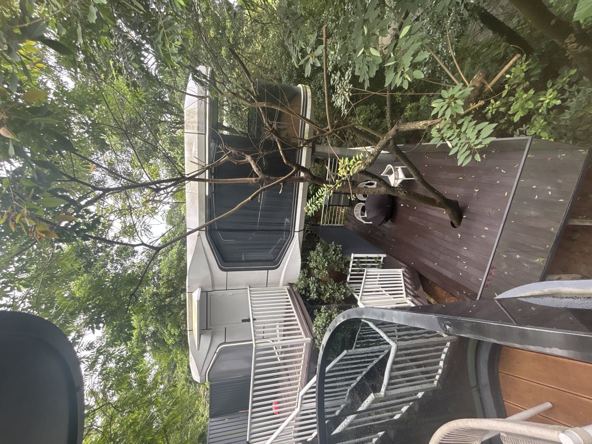 This image depicts a modern, elevated structure resembling a treehouse, nestled within a dense, green environment characteristic of a lush outdoor setting. The structure, with its white, curved facade and large, dark-tinted windows, appears to be suspended amongst the trees. A wooden deck, accessible by a winding staircase with white railings, is visible. Two white chairs and a black umbrella-like object are placed on the deck. The surrounding vegetation is abundant, with numerous trees and foliage creating a sense of seclusion. The overall impression is one of a unique, nature-integrated dwelling.