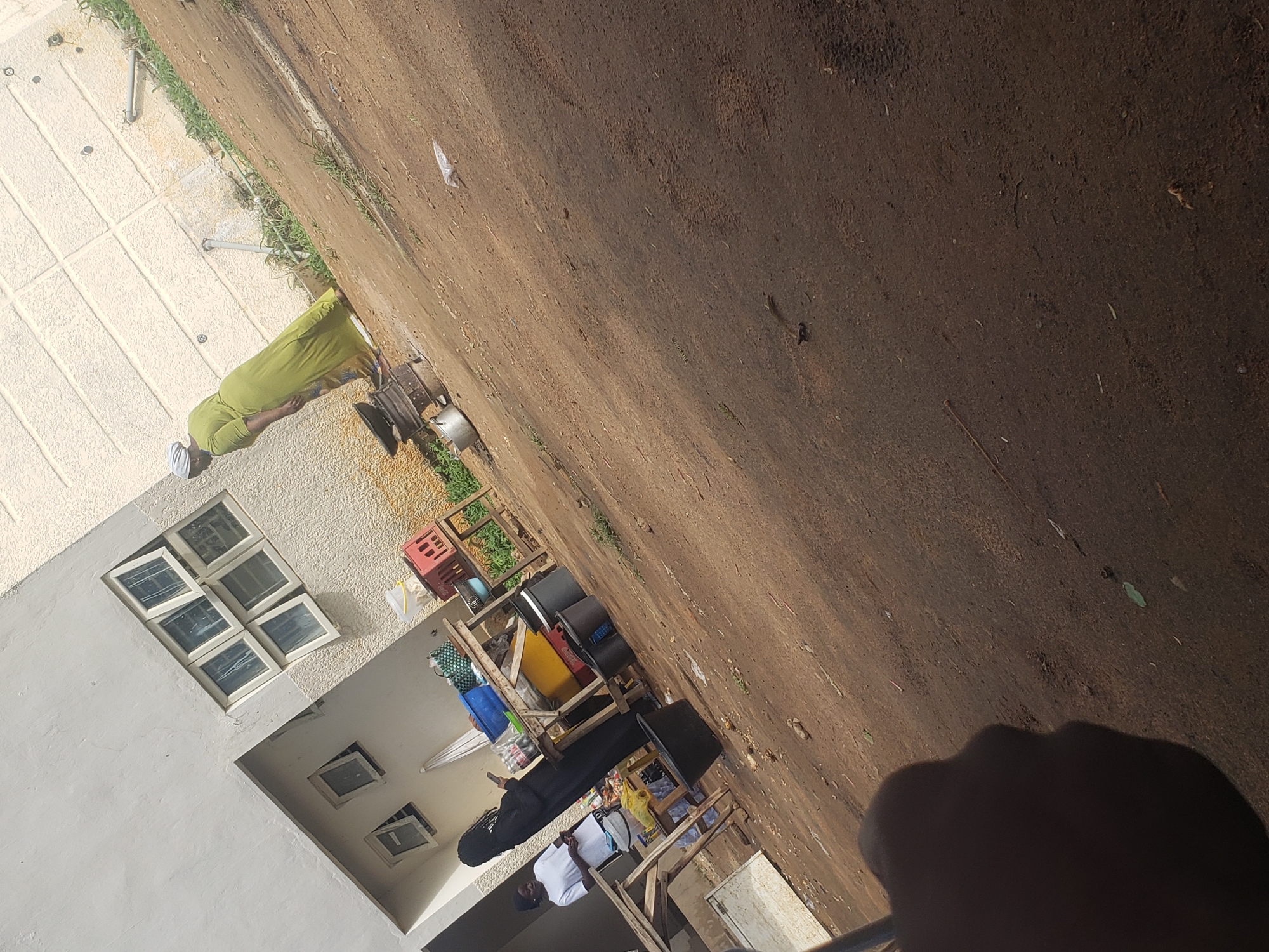 In an outdoor setting in Gumel, Nigeria, a woman in a lime green dress and headscarf stands near a simple metal stove and cooking pots. She appears to be looking down at something on the stove. To her left, a young woman in a white t-shirt and dark headscarf stands with her hands clasped, also looking downwards. Behind them, a makeshift market stall is set up with wooden tables displaying various items including baskets, containers, and crates. A large black bucket is also visible near the stall. The ground is dirt, with some scattered debris and sparse vegetation. In the background, a light-colored building with multiple windows is visible. The scene suggests a casual, everyday moment in a market or residential area.