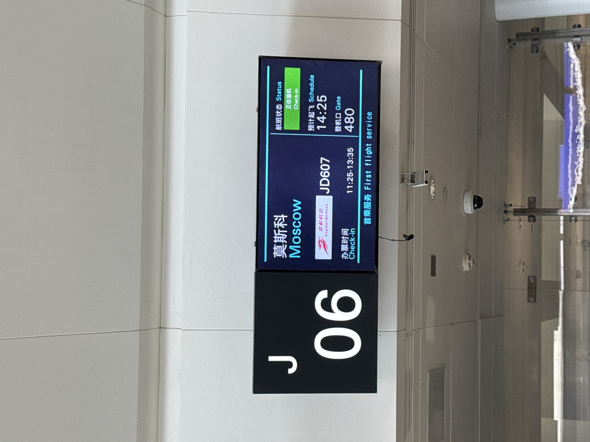 This is a medium shot of a digital display board at an airport in Xiaoshan District, China. The display board is divided into two sections. The left section is black and displays a large white "J" and "06". The right section is blue and displays flight information in both Chinese and English. The flight destination is Moscow, and the flight number is JD607. The status of the flight is "Check-in," and the scheduled departure time is 14:25. The gate number is 480. The display also indicates "First flight service." The background of the image is a white tiled wall. There are security cameras visible on the ceiling above the display board.