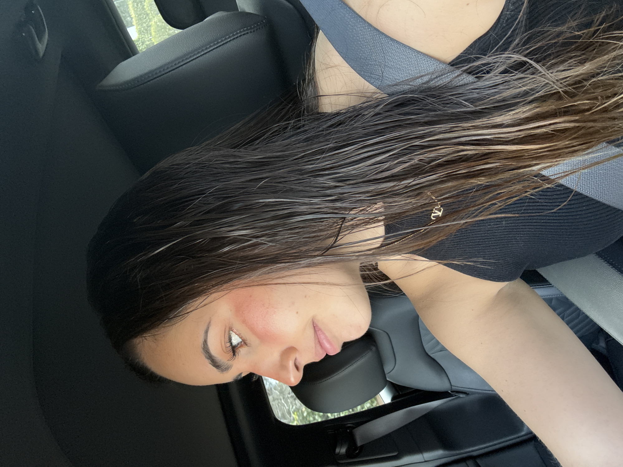 A young adult female is depicted seated in the driver's position of a vehicle in San Bernardino Tlaxcalancingo, Mexico. She is shown in profile, looking towards the left side of the frame. Her long, dark hair appears wet and is pulled back. She has a fair complexion with a distinct application of reddish blush across her upper cheeks, defined eyebrows, and visible eyelashes. Her lips are slightly parted. She is wearing a black ribbed tank top and a gold-colored necklace with a 'V' shaped pendant is visible on her chest. A grey seatbelt is properly fastened across her left shoulder and chest. The vehicle's interior features dark-colored, possibly leather, seating. Through the car windows, blurred green foliage is visible, suggesting an outdoor environment with significant vegetation.