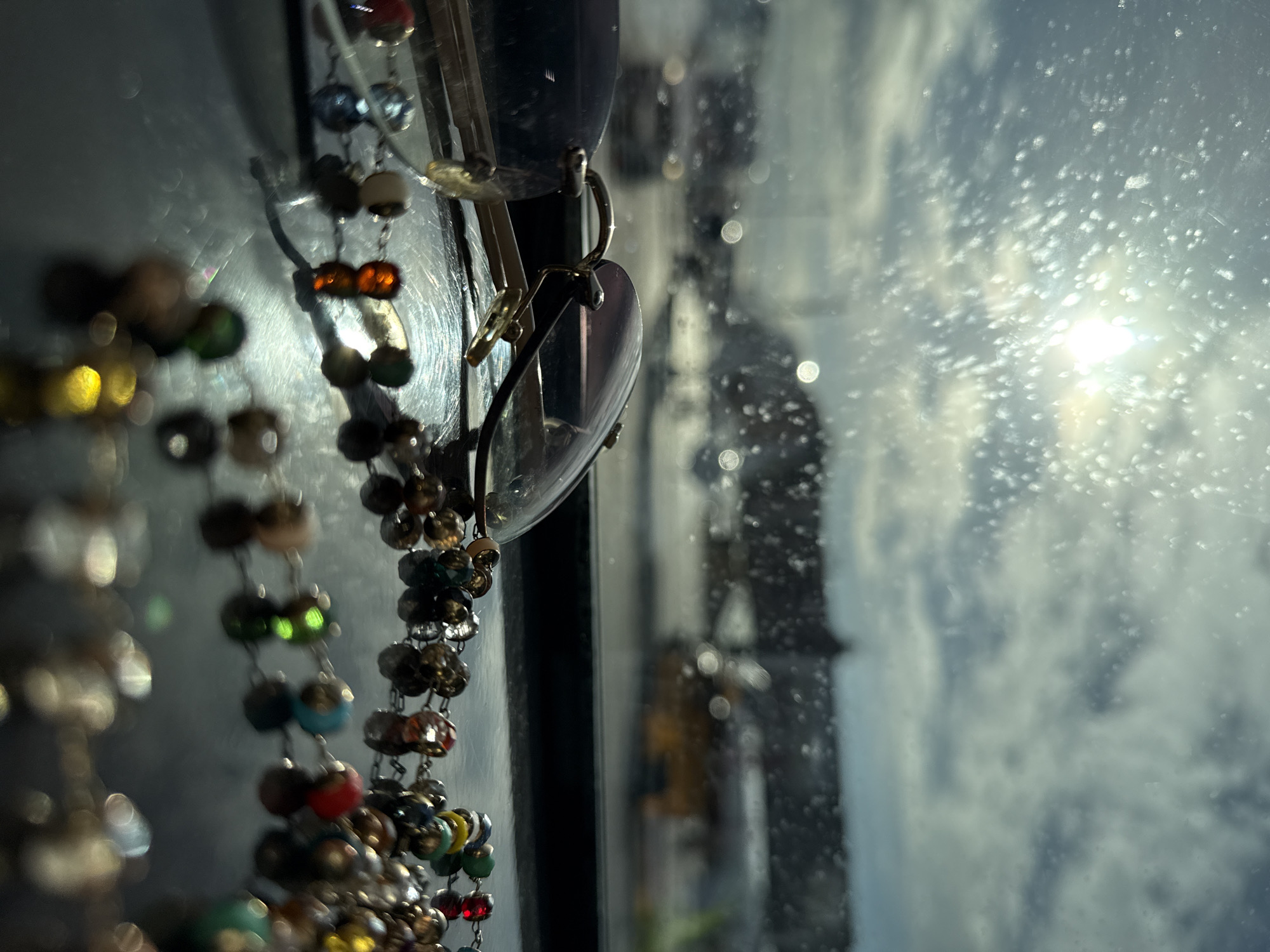 The image shows a close-up view through a dirty window with a cloudy sky and a bright sun outside. Hanging from the window frame are a pair of sunglasses and a colorful beaded chain. The location of the image is Khimki, Russia. There is no further information available from the image regarding people, subjects, objects, actions, or interactions.