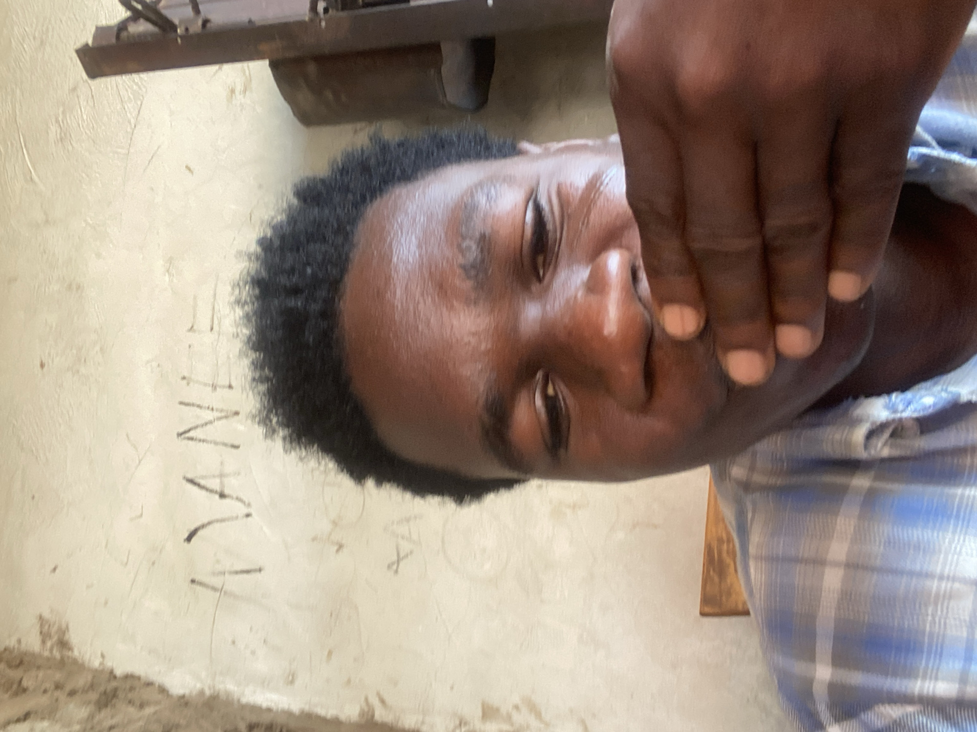 A close-up shot features a dark-skinned person lying on their side. The person has short, dark hair and is wearing a blue and white plaid shirt. Another dark-skinned hand is covering the person's mouth and nose. The background shows a wall with the name "NYANZI" scrawled on it. The location, Mararaba, Nigeria, is mentioned in the prompt and should be considered the setting of the image. There is machinery above the person.