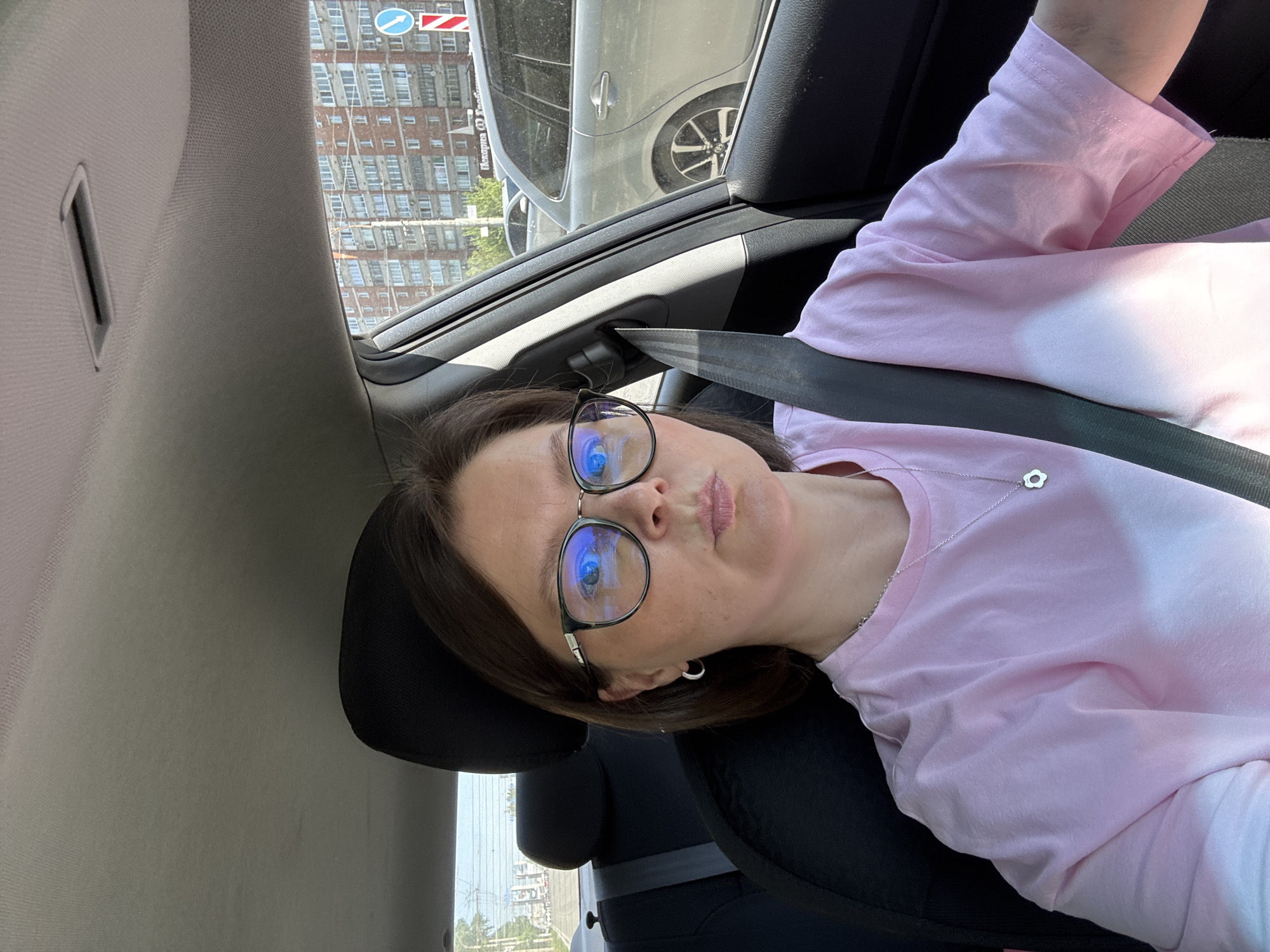 A person is in the passenger seat of a car. They are wearing a pink shirt, glasses, and a seatbelt. The reflection in their glasses suggests the scene is outdoors. The location is Izhevsk, Russia, identifiable by the street sign visible through the window of the car. Other cars and buildings are also visible through the window.