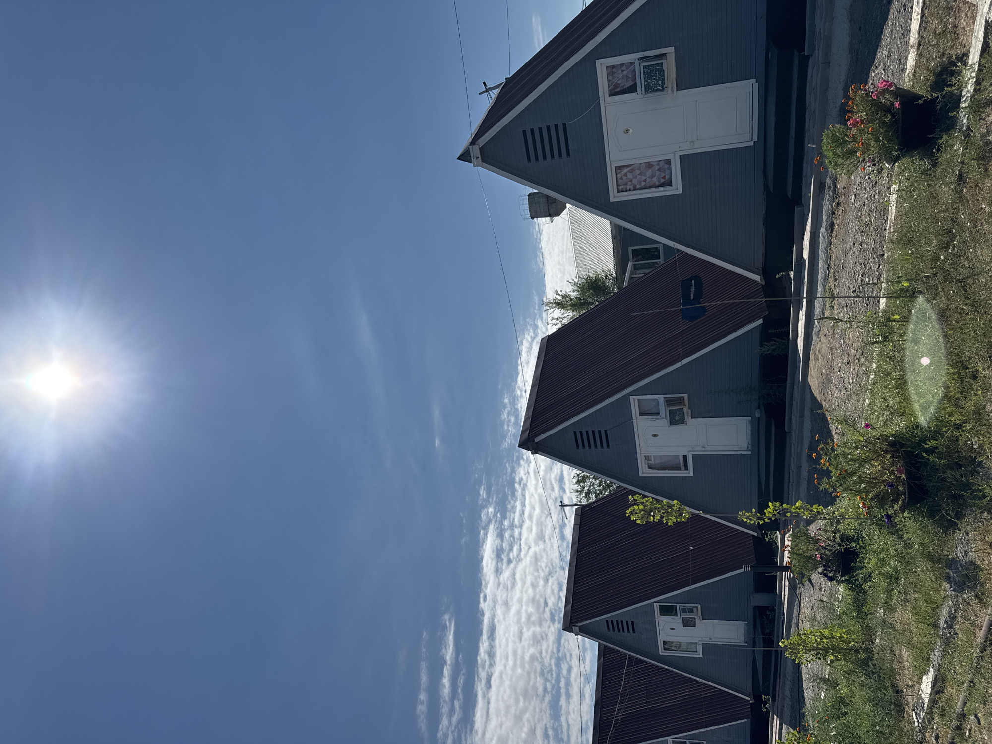 In Urdzhar, Kazakhstan, a row of identical triangular A-frame cabins with blue siding and dark red corrugated metal roofs are lined up under a bright blue sky with a strong sun and scattered white clouds. Each cabin features a white door, a small window with a patterned curtain, and an air conditioning unit installed below the window. In the foreground, lush green grass and potted plants with colorful flowers line the pathway in front of the cabins. The scene appears to be a peaceful outdoor setting, possibly a resort or a residential area.