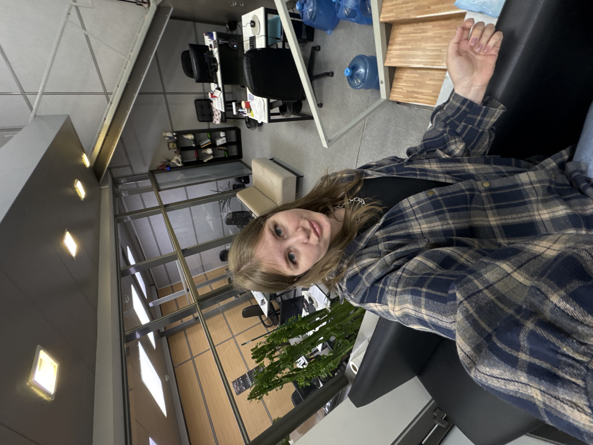 A young woman is captured in a selfie, looking directly at the camera with a neutral expression. She has long, light brown hair and is wearing a dark blue and beige plaid shirt over a black top, accessorized with a chain necklace. Her left hand, adorned with light-colored nail polish, rests on a black armrest, suggesting she is seated on a sofa or chair.
The setting appears to be a modern office or co-working space. In the background, multiple office desks with computer monitors and chairs are visible, along with papers and various office items. A tall, green, multi-branched plant stands prominently amidst the office furniture. Further back, a beige two-seater sofa is present.
The architecture includes a high ceiling with white panels and recessed square lights, along with large glass partitions or windows framed by metal supports. Some rectangular lights are mounted on a wall or structural column. To the woman's right, a water cooler with a large blue bottle is situated next to a light-colored, multi-shelf unit. Several blue plastic bags are also visible near the water cooler. The floor is a light, possibly tiled or concrete, surface.
The scene is lit by artificial indoor lighting. No discernible text is visible anywhere in the image. The overall impression is that of a functional and contemporary office environment located in Perm, Russia.
