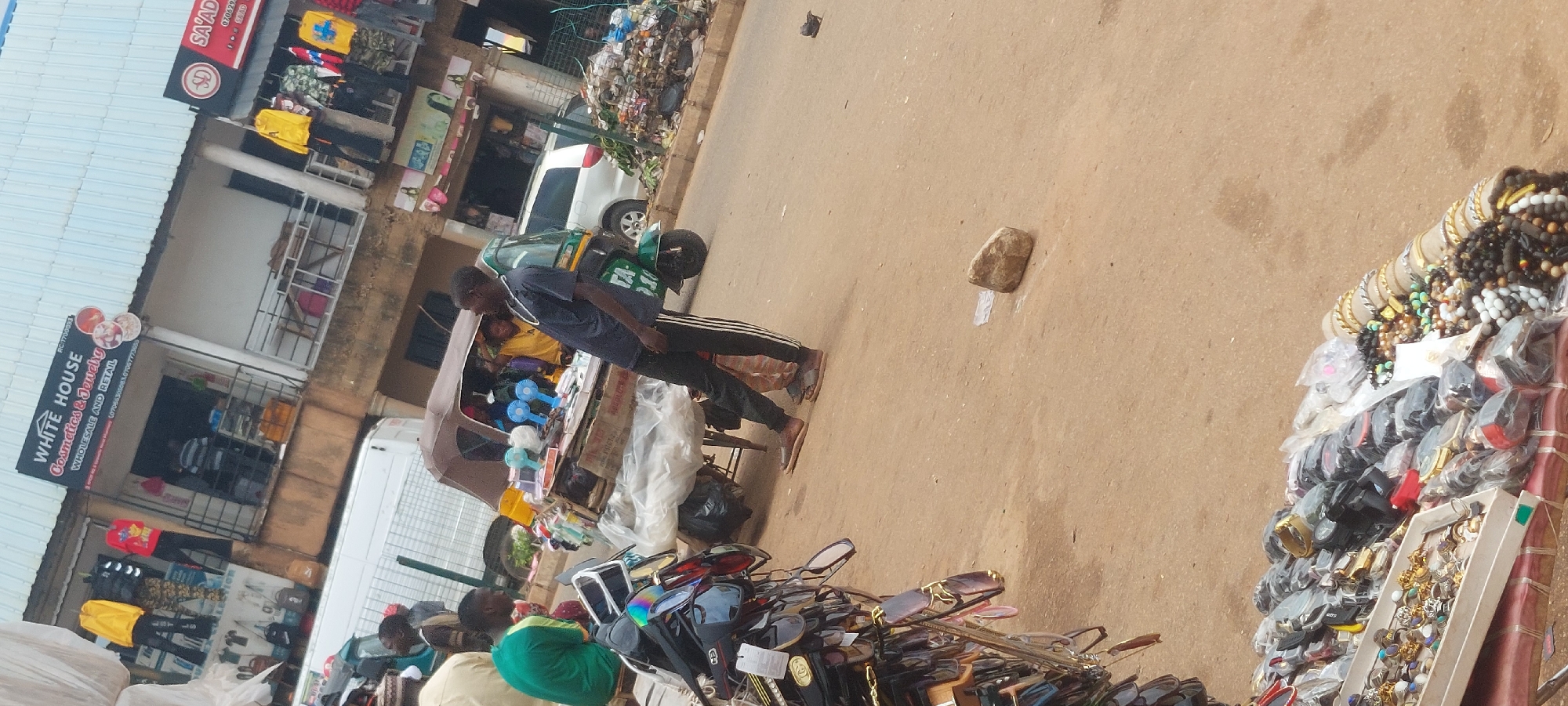 In an outdoor urban environment, a scene unfolds with a collection of stalls and a busy street. The focal point is a man standing to the right, wearing a dark blue t-shirt with white stripes on the sleeves and black pants with white stripes down the sides. He is looking to his right and appears to be interacting with a vendor. In front of him, a cart is laden with various items including sunglasses on display, small fans, and other assorted goods. Behind the cart, a three-wheeled vehicle, possibly an auto-rickshaw, is parked. The background shows a building with shops, one of which is labeled "WHITE HOUSE Cosmetics & Jewelry". Further to the right, another shop sign is visible with "SD SA'AD". The ground is a dirt or gravel surface. There is a pile of garbage visible on the right side of the image. Several people are present, including a person in a green shirt to the left of the cart and others partially visible within the shops and the vehicle. The lighting suggests it is daytime, with some shadows indicating overhead structures or the sun's position.