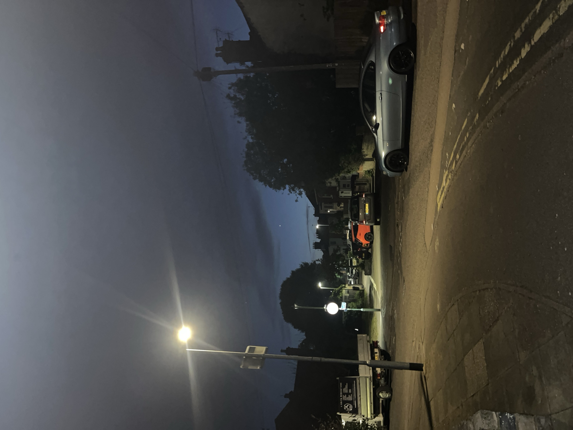 The image depicts a street scene in East Suffolk, United Kingdom at dusk or night. The photograph is taken vertically. A light-colored car, possibly silver or light blue, is parked on the right side of the road. The street has double yellow lines on the asphalt and a paved sidewalk to the right of the road. Further down the road, there is an orange car. A white street light illuminates the foreground. A tall tree is beside the street. A truck is parked next to the street light. Houses and buildings are seen in the distance. The sky is dark.