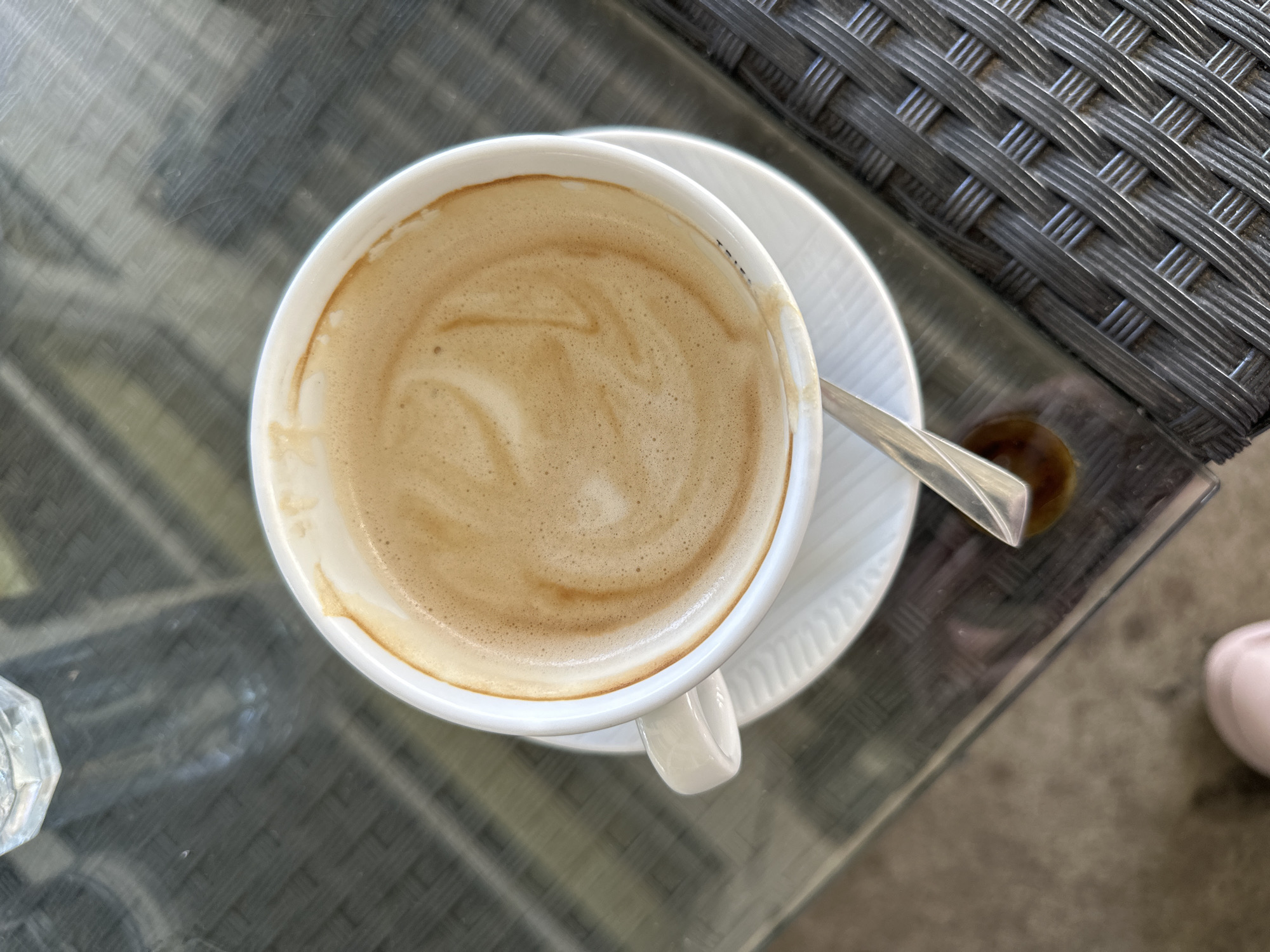 A top-down view captures a white ceramic cup of coffee with foam art on a saucer, placed on a glass-topped table with a woven rattan edge. A silver spoon rests partially in the cup, with a small drip of coffee beside it. In the background, a clear glass is partially visible. The scene appears to be outdoors, possibly on a patio, with diffused light suggesting daylight. The overall impression is one of a relaxed moment, perhaps at a cafe in Grad Rijeka, Croatia.