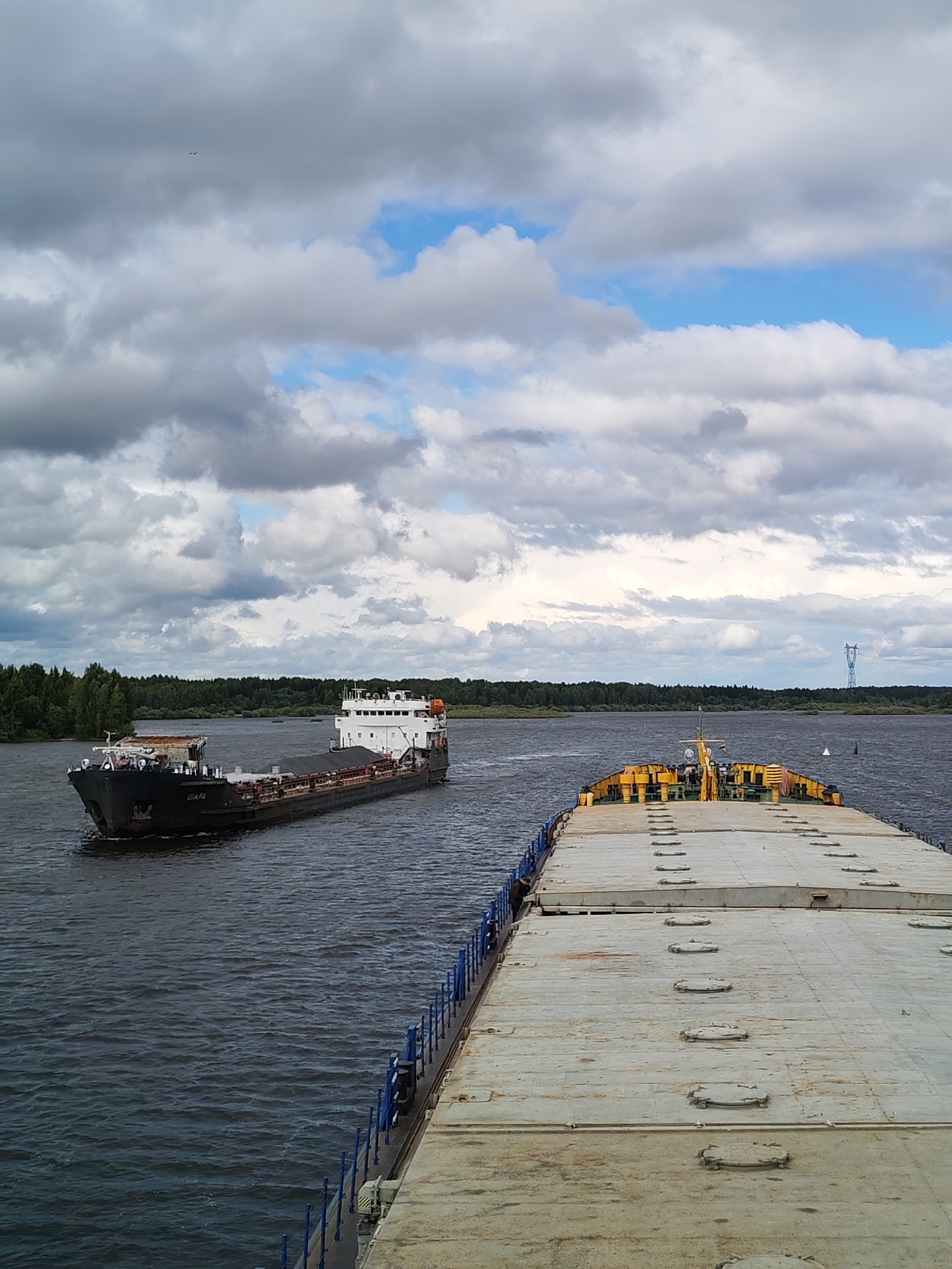 The image shows a wide river with two barges. In the foreground, a flat-topped barge is heading away from the viewer, its deck covered with rectangular hatches. The barge has blue railings along its sides. In the middle ground, a second, darker barge with a white superstructure is moving in the opposite direction. This barge appears to be carrying cargo. The riverbanks are lined with dense green trees. The sky is cloudy with patches of blue sky visible. A power line tower is visible in the distance on the right side of the frame. The scene is likely located on the Volga River near Sud'bitsy, Russia, as indicated by the presence of barges and the characteristic landscape.