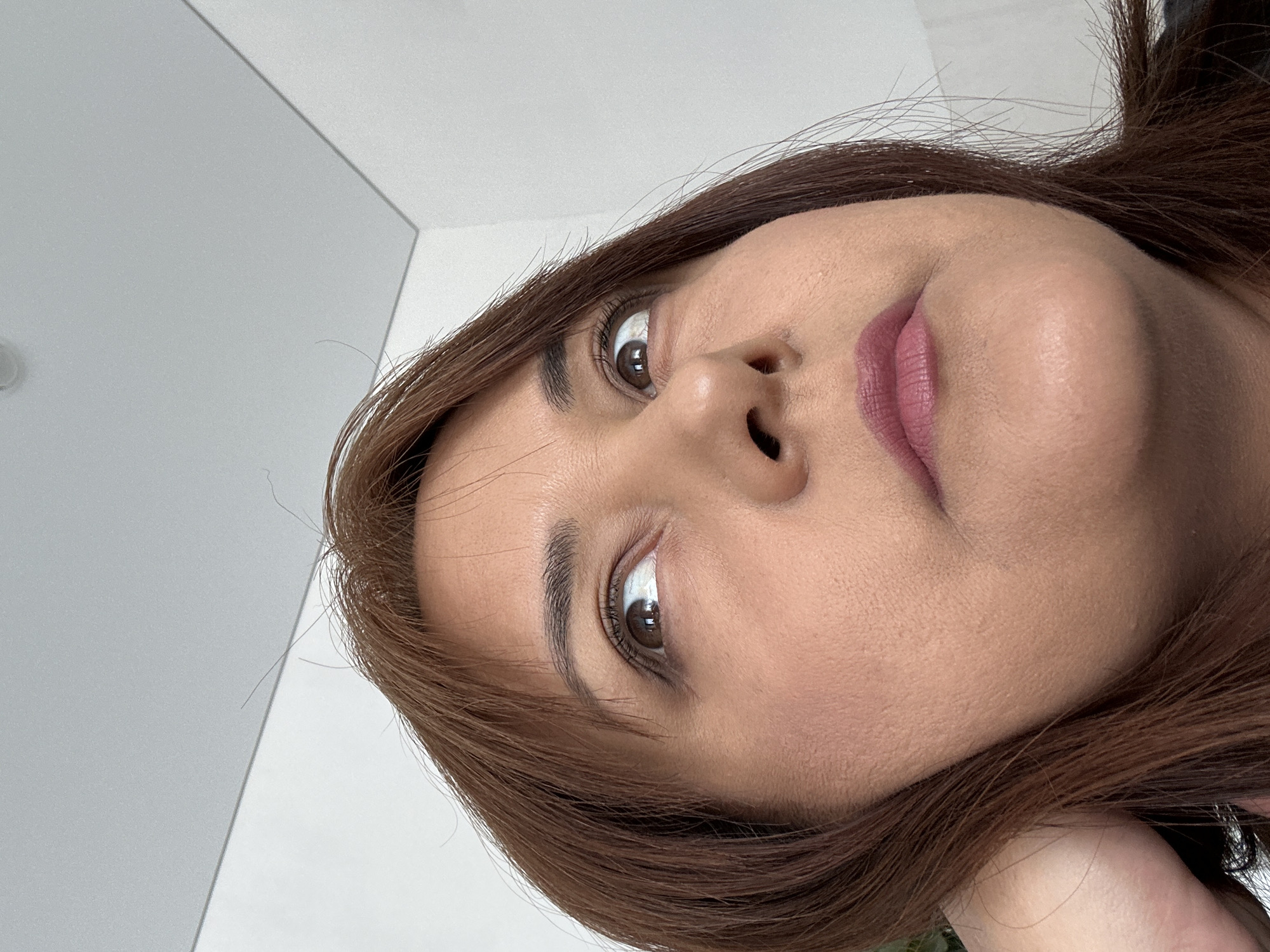 A close-up, low-angle shot of a woman's face is presented. The woman has light brown hair and brown eyes. Her skin appears fair, and she is wearing lipstick. The background features a white wall. The location is unknown.