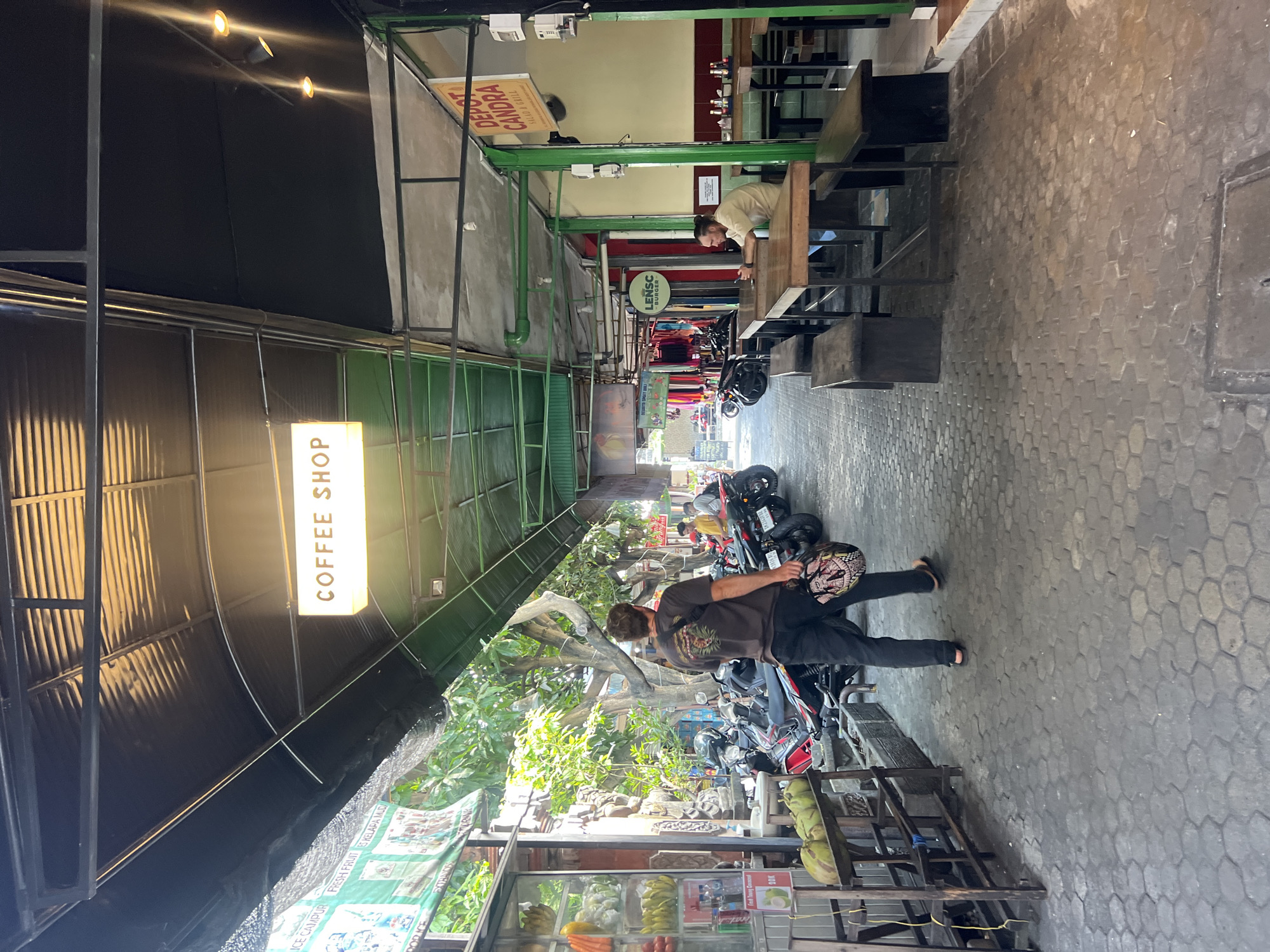 The image shows a street scene in Kuta, Indonesia, with a covered walkway over a paved area. A man in a dark t-shirt and black pants is walking away from the camera, carrying a helmet. He is on the left side of the walkway, which is lined with parked motorcycles. To the right, there are several tables and benches, suggesting an outdoor dining area. A sign above the tables reads "DEPOT CANDRA SALAD & GRILL." Another sign in the distance reads "COFFEE SHOP." The scene appears to be in a commercial or tourist area, with shops and food establishments. Trees and foliage are visible in the background, indicating a tropical environment. The ground is paved with hexagonal blocks.
