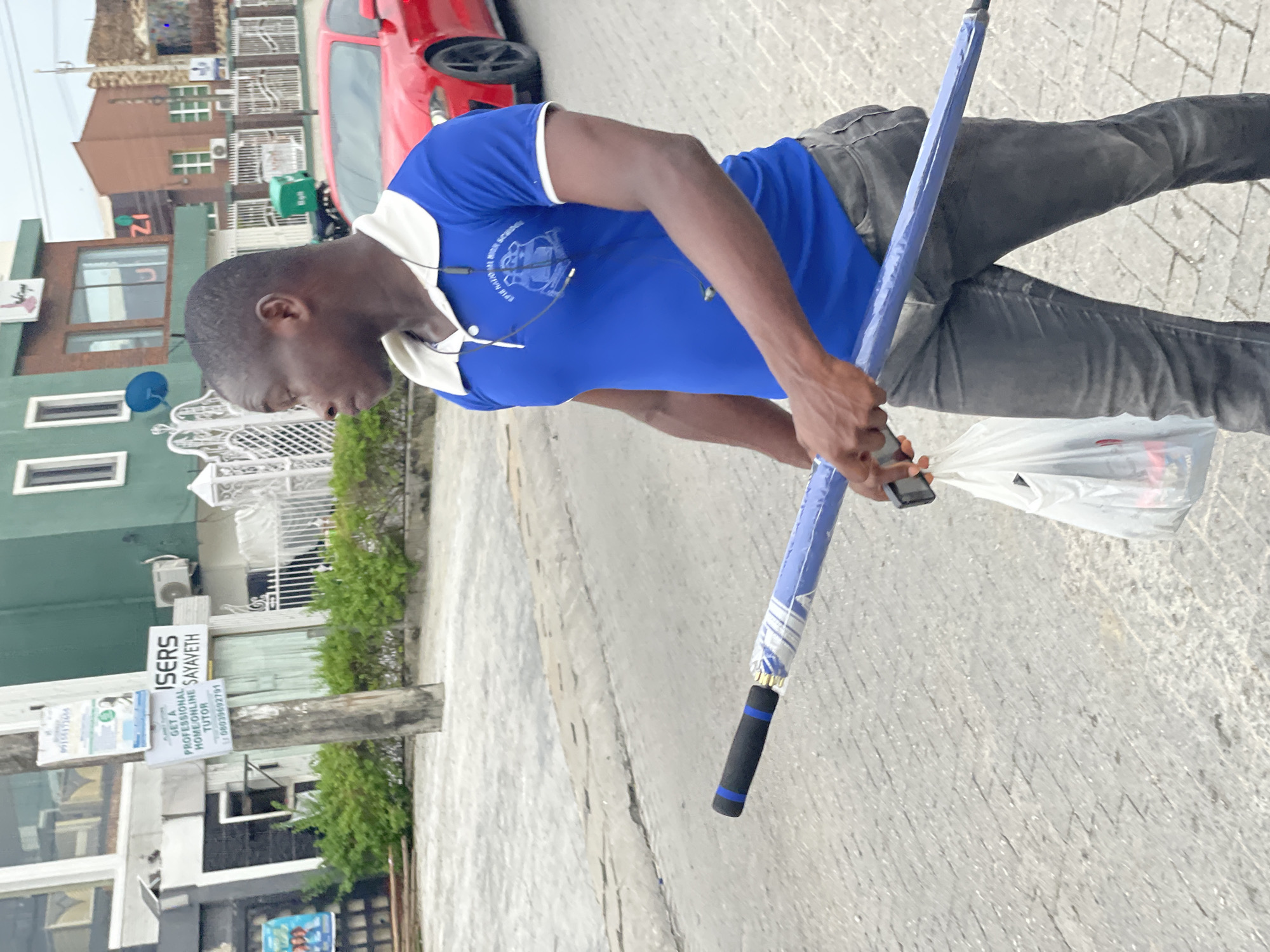 A Black man is walking on a paved street in Moba, Nigeria. He is wearing a blue polo shirt with a white collar and a school emblem, dark grey jeans, and appears to be in his 20s or 30s. In his right hand, he is carrying a blue umbrella with white accents, and in his left hand, he is holding a white plastic bag. Behind him, a red sports car is parked on the street, and buildings with signs are visible in the background, including one with the word "ISERS" and another with "ZI". The environment appears to be a commercial or residential area within the city.
