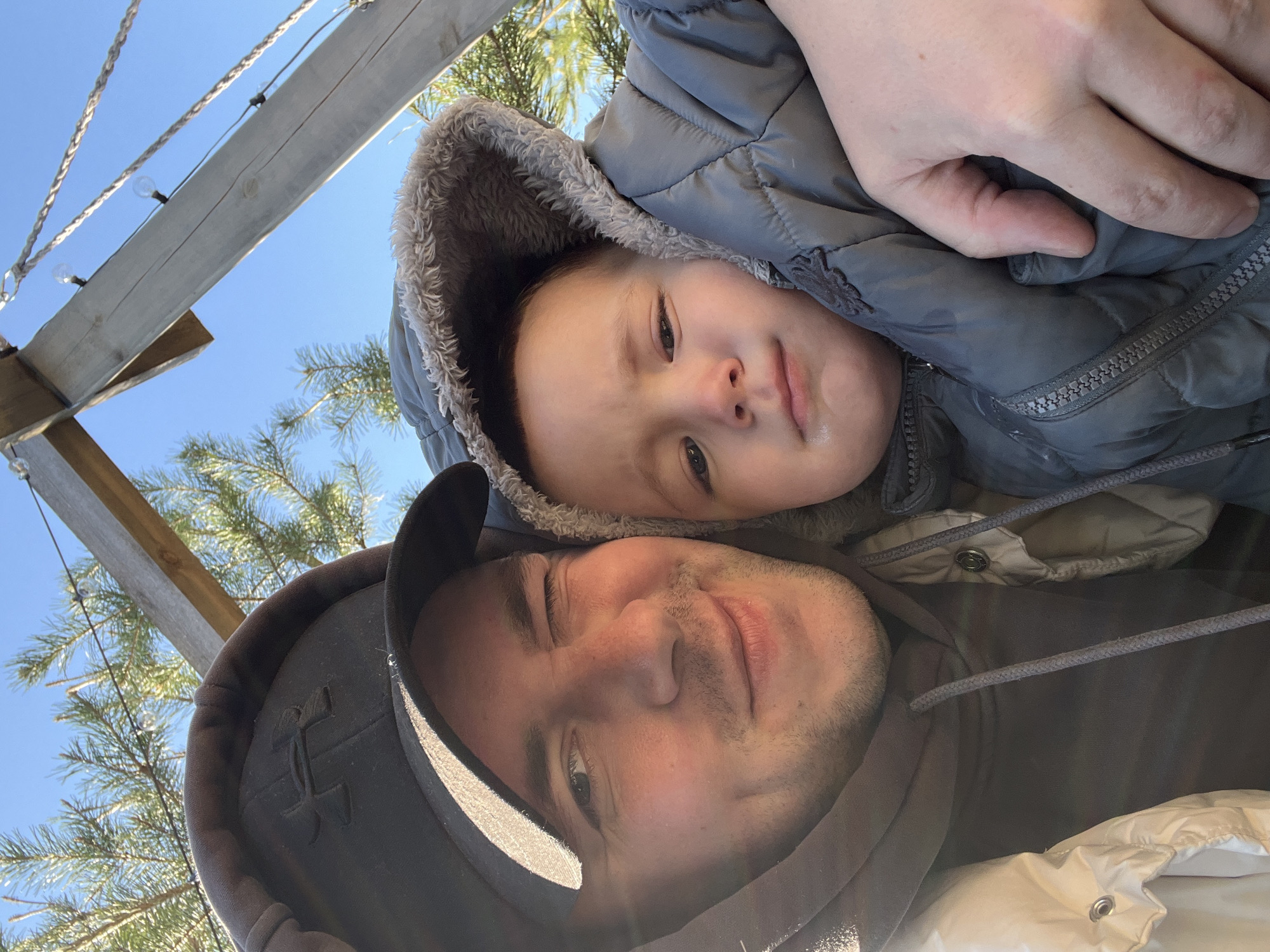 A low-angle selfie, likely taken in Litvinovo, Russia, shows an adult male and a child, possibly in an outdoor setting. The adult male wears a dark-colored hooded sweatshirt and a baseball cap. The child is bundled in a hooded grey puffer jacket with a fur-trimmed hood. The child is close to the adult, possibly being held. The background includes evergreen trees, what appears to be a wooden structure with string lights, and a clear blue sky.