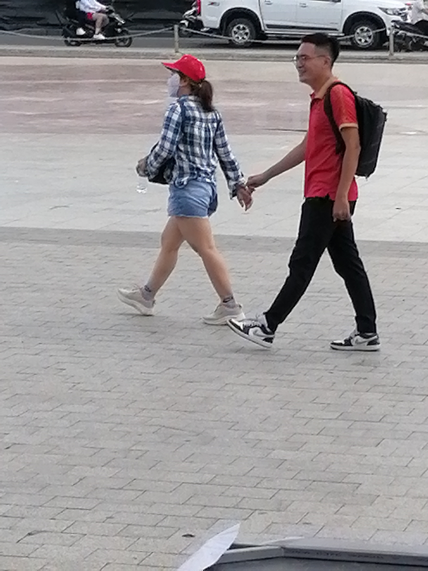 A man and a woman walk hand-in-hand across a paved plaza. The woman is on the left, wearing a blue and white plaid shirt, denim shorts, a red baseball cap, and a face mask. She carries a clear plastic bottle in her left hand. The man, on the right, wears a red t-shirt, black pants, and a black backpack. He has on glasses. They are walking on a stone-tiled surface with a white line marking a boundary in the foreground. In the background, a white pickup truck is parked, and a motorcycle is visible. The scene appears to be in Da Lat, Vietnam.