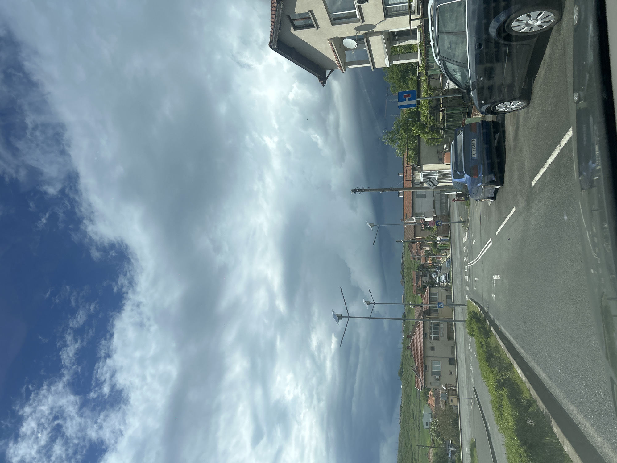 The image depicts a street scene, likely taken from inside a vehicle. The sky is dominated by dramatic cloud formations, transitioning from patchy blue sky with cumulus clouds to a dark, ominous storm cloud mass. A road stretches into the distance, lined with buildings and green vegetation on either side. The buildings appear to be residential, with red-tiled roofs and light-colored facades. Streetlights are visible along the road. Several parked cars are present, including a dark-colored van and a blue car with license plate "A 3156 HT" visible. The location is likely in the city of Burgas, Bulgaria, based on the license plate of the blue car.