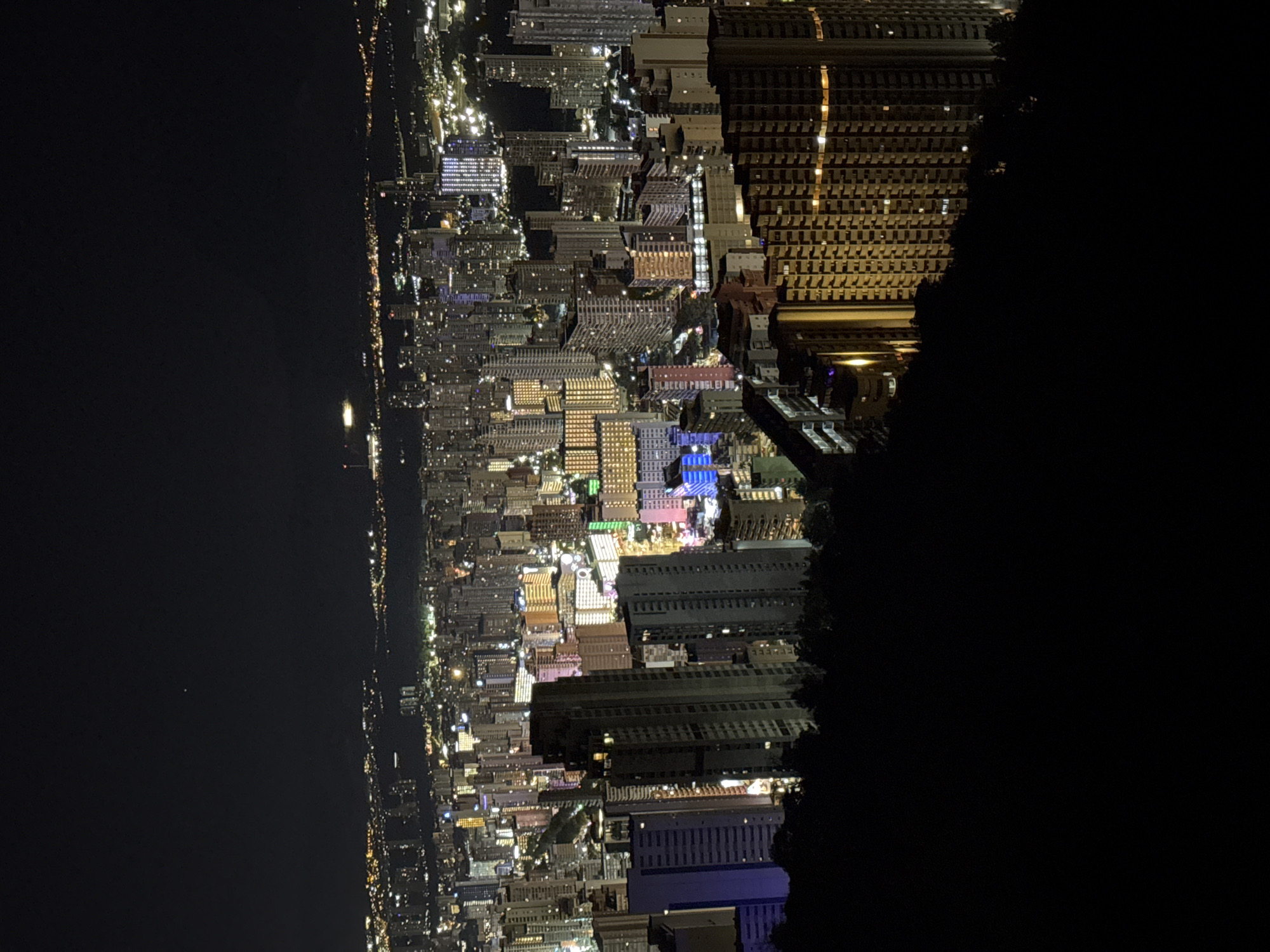 This is a night-time aerial photograph of the city of Benidorm, Spain. The foreground is dominated by a dark, silhouetted hillside, covered in trees. Beyond this, the city sprawls out with numerous high-rise buildings illuminated by artificial lights. The buildings vary in height and architectural style, with many featuring balconies and window lights. Some buildings are brightly lit with colored lights, particularly in hues of pink, blue, and yellow. In the background, the distant lights of more buildings and possibly a coastline are visible, stretching towards a dark, starless sky. The overall impression is of a densely populated urban area at night.