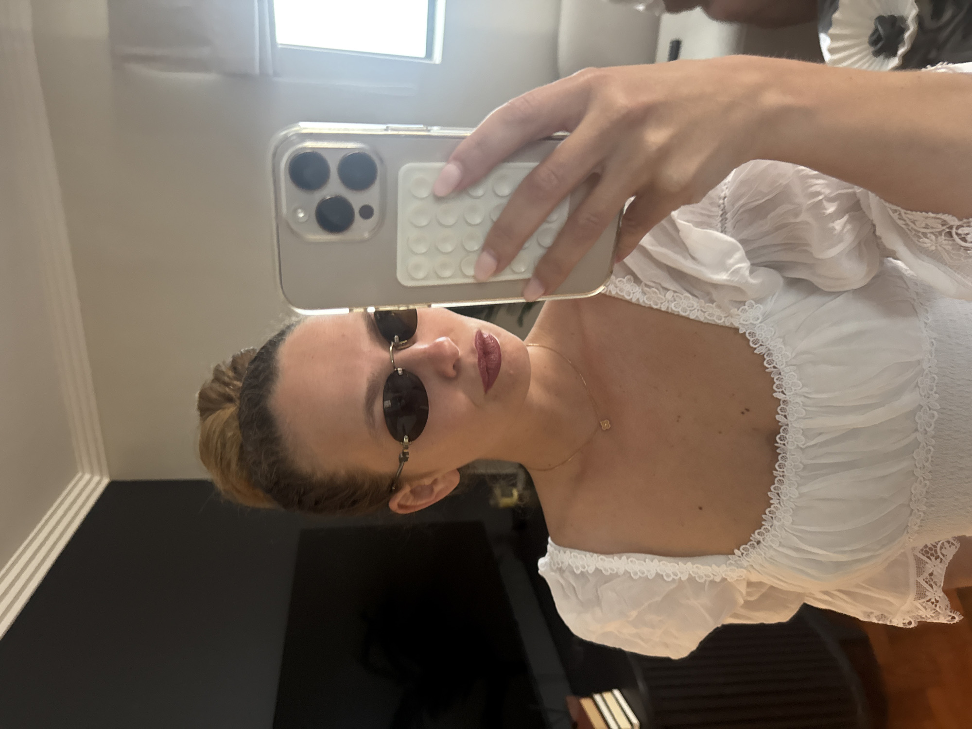A woman is taking a selfie in a room with neutral-colored walls, a window, and a dark television. She is wearing a white top with lace details and round sunglasses. She is holding a smartphone with a silver case and a white accessory on the back. There is no indication of the location being in Cape Town, South Africa.