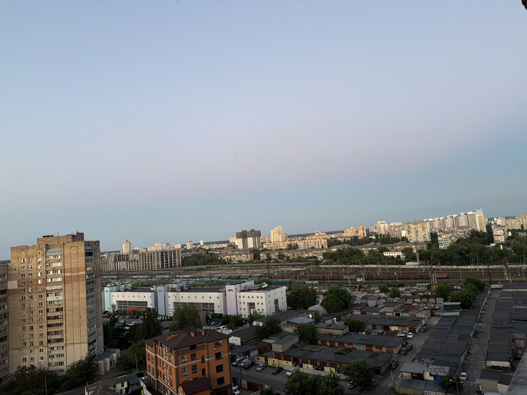 This is a high-angle, wide shot photograph of an urban landscape in Kyiv, Ukraine, likely during the late afternoon or early evening given the soft lighting. The foreground features a cluster of low-rise garages, many with corrugated metal roofs, interspersed with trees and parked cars. Behind the garages, a large industrial or commercial complex with white and blue buildings is visible, along with train tracks and several train cars. Further in the distance, a dense residential area with mid-rise apartment buildings stretches across the horizon, punctuated by taller, modern high-rises. The sky is clear and light blue with a hint of haze. The scene depicts a typical mixed-use area of a large city, combining residential, industrial, and transportation infrastructure. No people are visible in the frame.