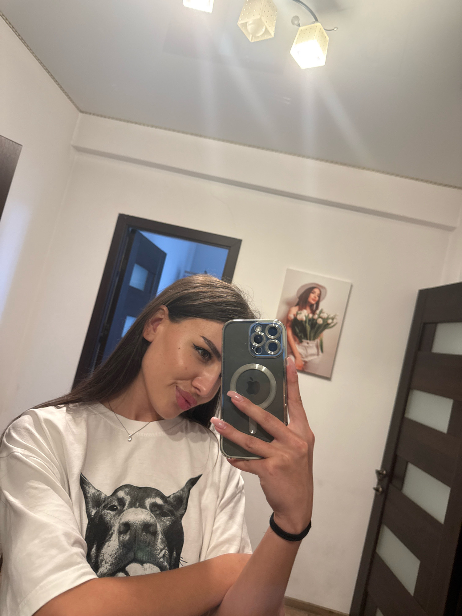 A young woman with long, dark hair takes a mirror selfie in a well-lit room. She is wearing a white t-shirt with a large, black and white graphic of a dog's face. She has on a delicate silver necklace. In her right hand, she holds a smartphone, angled to capture her reflection. The background shows a white wall with a framed picture of a woman holding a bouquet of flowers, a dark brown door with frosted glass panels, and a partially open door revealing a blue-tinted interior. The ceiling has modern light fixtures. The overall impression is of an indoor domestic setting.