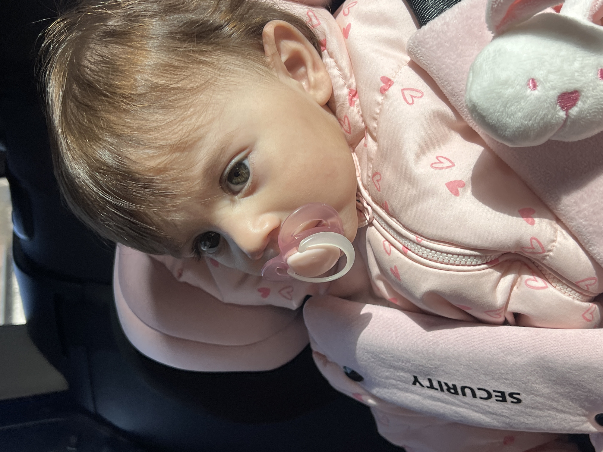 A close-up shows a baby lying in a car seat. The baby has light brown hair, fair skin, and brown eyes. The baby is wearing a pink jacket with small heart patterns and has a pacifier in its mouth. Next to the baby is a white stuffed bunny toy. The side of the car seat reads "SECURITY" in black letters. The environment suggests an indoor setting, likely a car, with dark upholstery visible in the background. There is no indication of the location being Elena, Bulgaria, within the provided image.