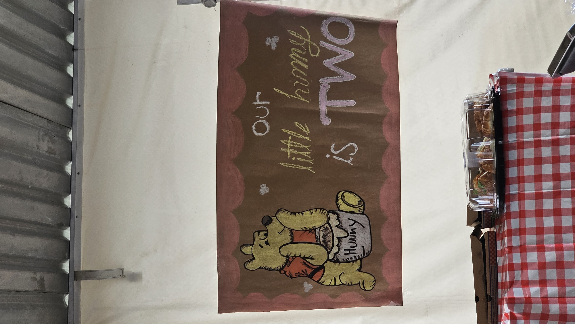 A hand-painted birthday banner hangs vertically. It depicts Winnie the Pooh lying on his back. The banner, painted on brown paper, is bordered with a scalloped edge in pink. The words "Our little Hunny is TWO" are written in a light pink, on the banner. To the left of the banner, there is a corrugated metal surface. To the right of the banner, there is a red and white checkered fabric. A clear plastic container holding croissants is also visible. The location is San Bernardino Tlaxcalancingo, Mexico.