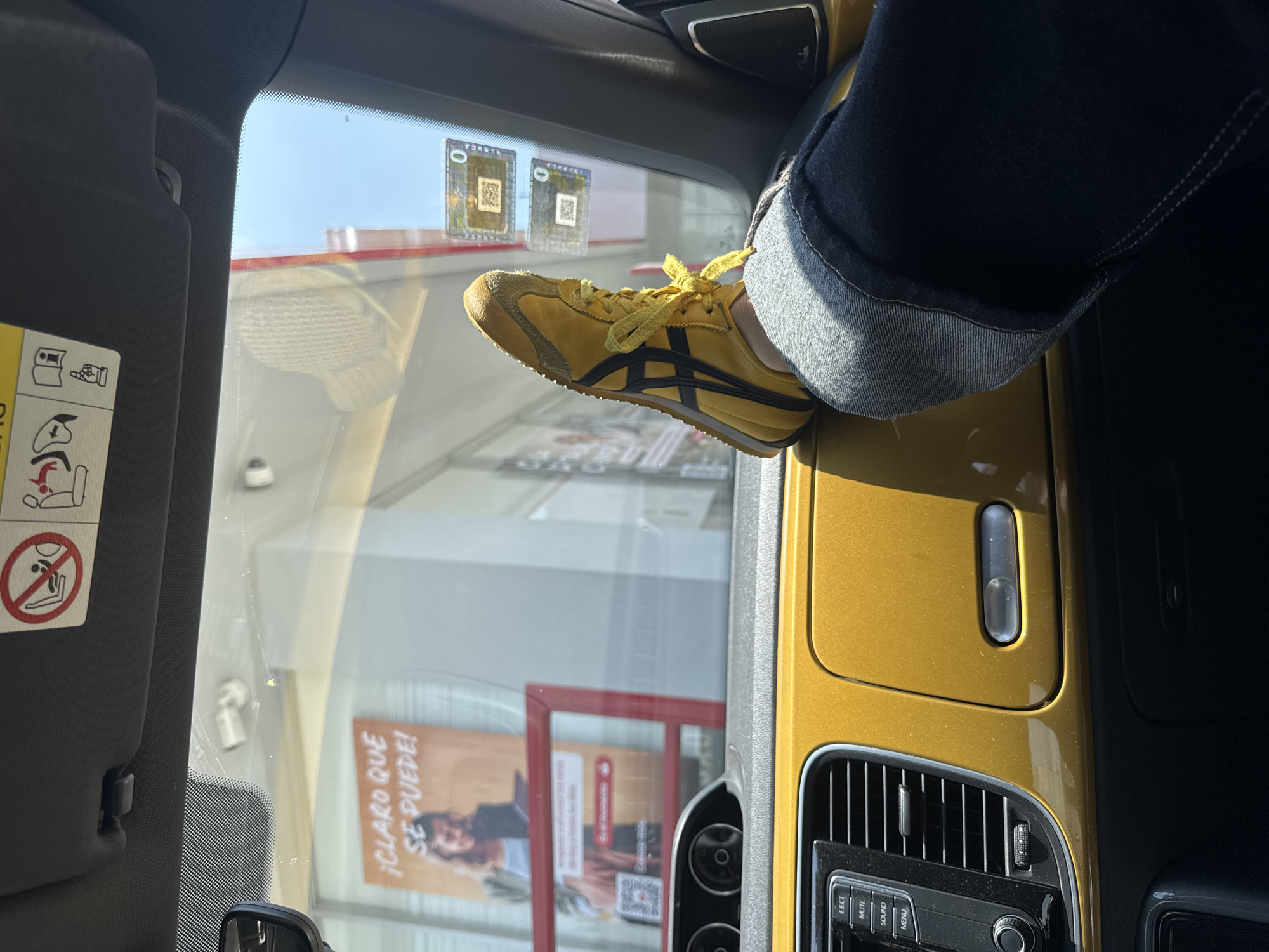 Inside the passenger side of a gold-colored vehicle, an individual wearing blue jeans with rolled-up cuffs has their left foot, adorned with a yellow Onitsuka Tiger shoe with black stripes, resting on the dashboard. The car appears to be parked or stationary. Visible through the windshield is the exterior of a commercial establishment in Puebla, Mexico. Window stickers are affixed to the upper portion of the windshield. The sun visor on the driver's side bears warning labels. The vehicle's dashboard features air vents, a stereo system, and other console elements.