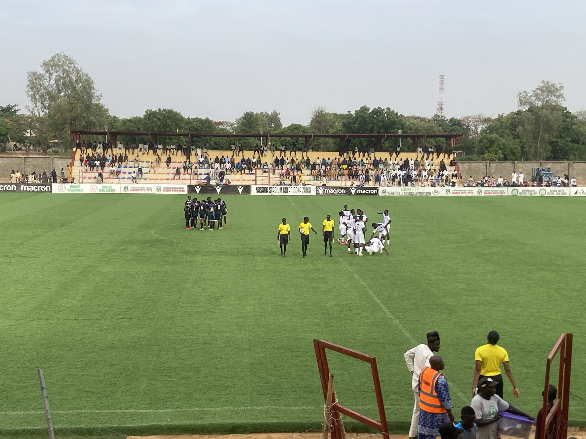 The image shows a soccer game at Nasara Stadium Kofar Doka Zaria, in Tudun Wada, Nigeria. Two teams, one in dark blue and one in white, are on the field with referees in yellow. Some players from the white team are interacting on the ground. A stand is visible in the background with spectators. In the foreground, individuals in varying attire, including a person in an orange safety vest, are standing near a fence. Logos and text are visible on the advertising boards surrounding the field, including "Macron", "Kaduna State Ministry of Sports Development", and "Nasara Stadium Kofar Doka Zaria."