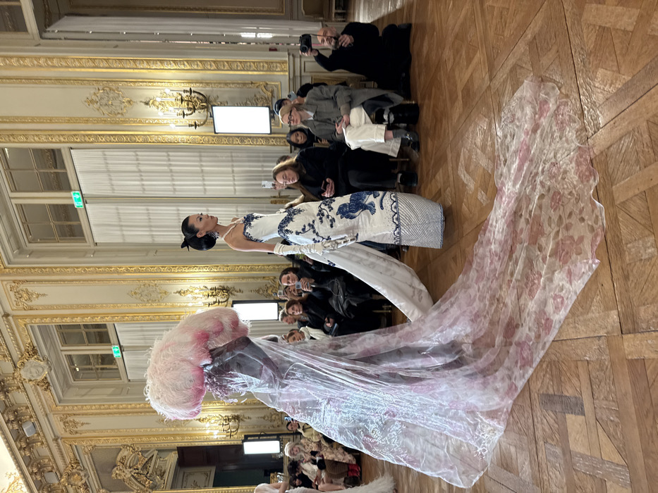 A fashion show presents two models. One wears a white and blue dress with intricate patterns and a long white train. The other is draped in a translucent, rose-patterned fabric, topped with a large, pink, feathery headpiece. The audience is seated on either side of the runway, capturing the scene with cameras. The venue is ornate with golden detailing, large windows, and a parquet floor.