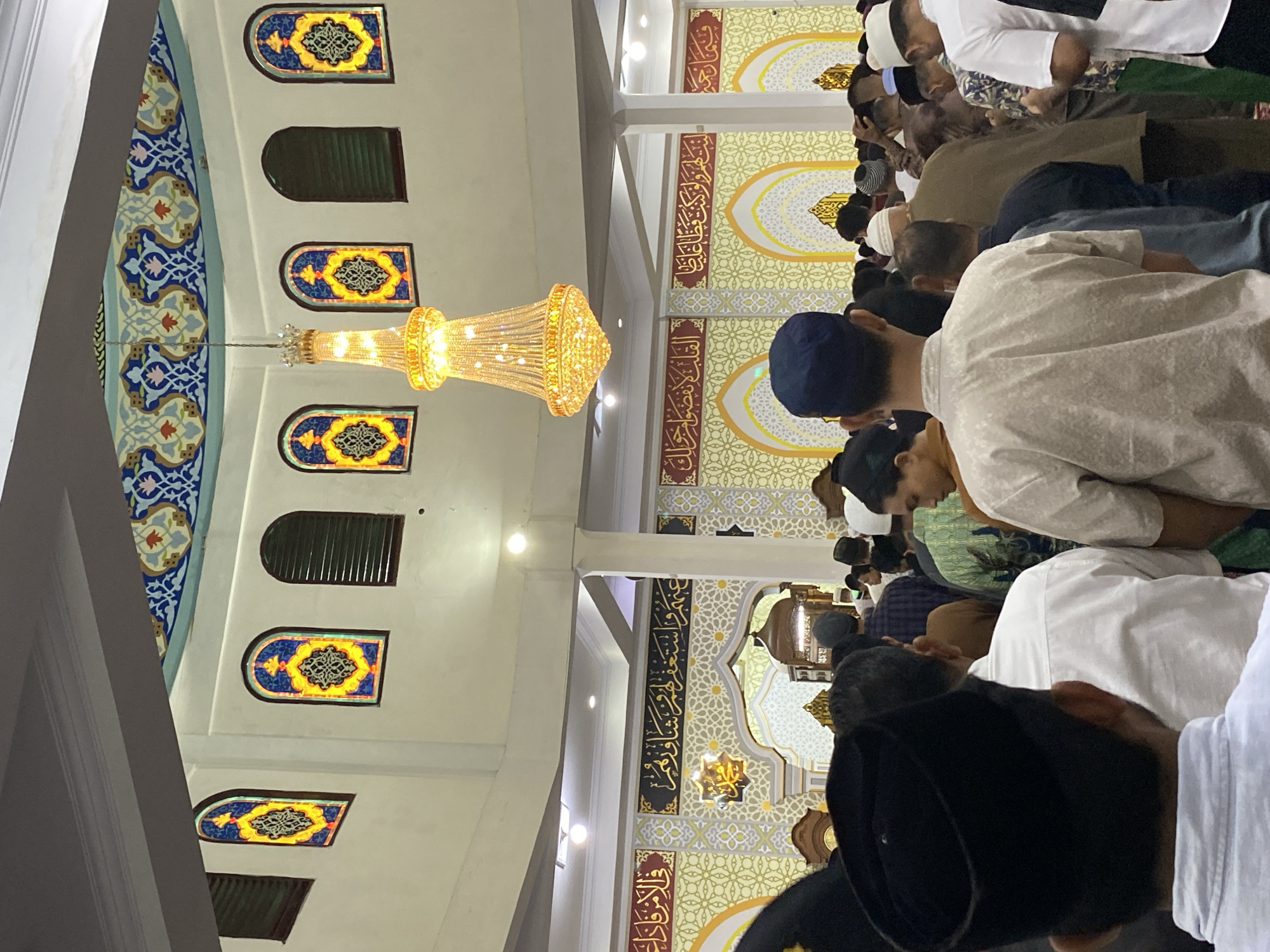 This is a photograph taken inside a mosque, likely in Lhokseumawe, Indonesia, showing a gathering of men in prayer. The interior features intricate Islamic calligraphy on the walls, arched doorways, and decorative stained-glass windows in arched frames. A large, ornate chandelier hangs from the ceiling, illuminated and casting a warm glow. The men, many wearing skullcaps, are seen from behind, facing a direction that suggests they are engaged in congregational prayer. Some individuals are dressed in traditional Islamic attire, while others wear more modern clothing. The atmosphere appears to be solemn and devout, typical of a religious service.