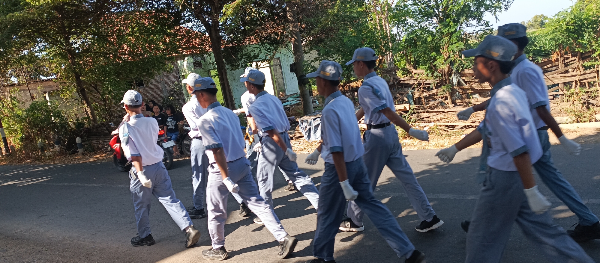 In Ketowan, Indonesia, a group of students wearing light blue shirts, gray pants, and gray caps walk in formation on an asphalt road. They are wearing white gloves and black shoes. A red motorcycle is parked on the side of the road behind some of the students. Several onlookers, including a few females wearing black shirts, are visible behind the motorcycle. The background consists of trees, a fence made of wood, and some buildings. The sun is shining, casting shadows on the road.