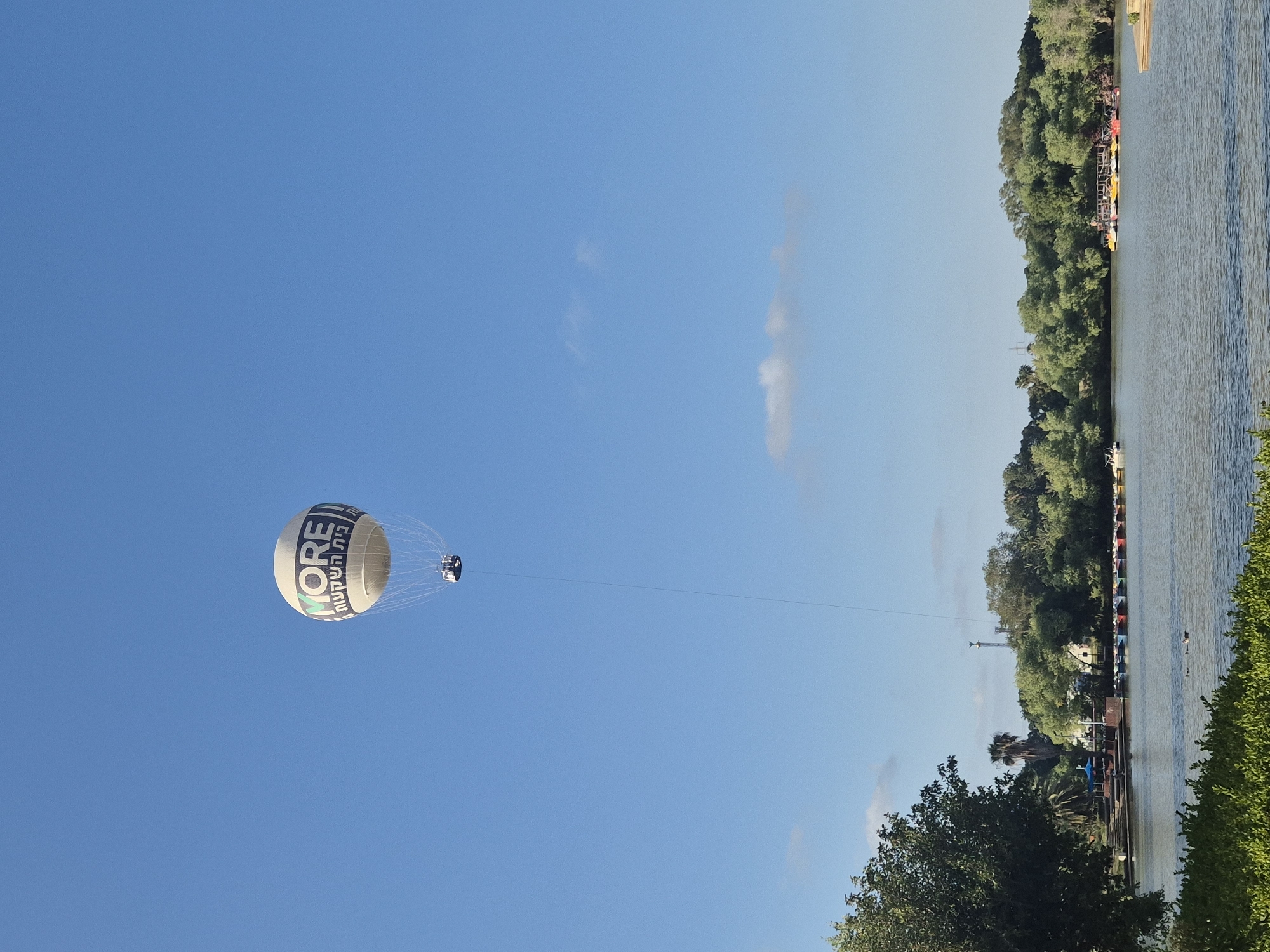 This daytime image, set in Tel Aviv, Israel, presents a vibrant recreational scene featuring a large, tethered hot air balloon against a bright, clear blue sky. The balloon, predominantly white with a light base, is emblazoned with the English word "MORE" in prominent dark letters, and beneath it, the Hebrew text "בית השקעות" (Beit Hashka'ot), which translates to "Investment House." A small gondola or basket, likely carrying passengers, hangs directly beneath the balloon, suspended by numerous thin lines. A thick tether extends downwards from the gondola, indicating it is anchored, suggesting a static or controlled ascent attraction.
The right side of the image features a calm body of water, likely a lake or river, stretching towards the background. Its far bank is densely covered with lush green trees and foliage. Along this tree-lined bank, a dock or pier is visible, where several colorful small boats, appearing to be kayaks or pedal boats, are lined up, suggesting water-based recreational activities. More green trees and bushes frame the lower right corner of the foreground. The sky is mostly devoid of clouds, apart from a few very faint wisps, indicating fair and sunny weather. The overall impression is one of leisure and outdoor enjoyment in a well-maintained park environment.