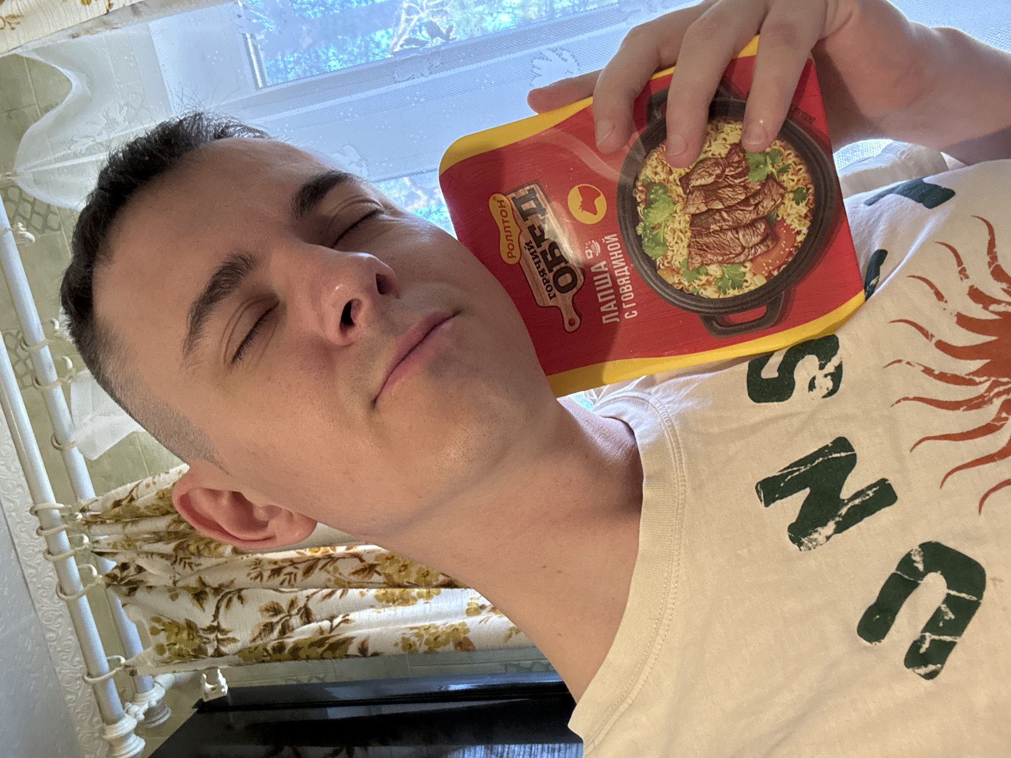 A fair-skinned male with short dark hair is photographed in a tilted, close-up shot. His eyes are closed and a packet of instant noodles rests against his face. The noodle packet is red and yellow, displaying an image of noodles with beef. The subject wears a white t-shirt with a large graphic print of the word "SUN" in a stylized font, with the letters appearing to be green.
The environment includes a window behind him, fitted with a white curtain. To the left, floral print curtains hang over what appears to be a radiator. The scene likely takes place indoors, perhaps in a residential setting. No specific location in Chuprino, Russia can be determined from the image.
